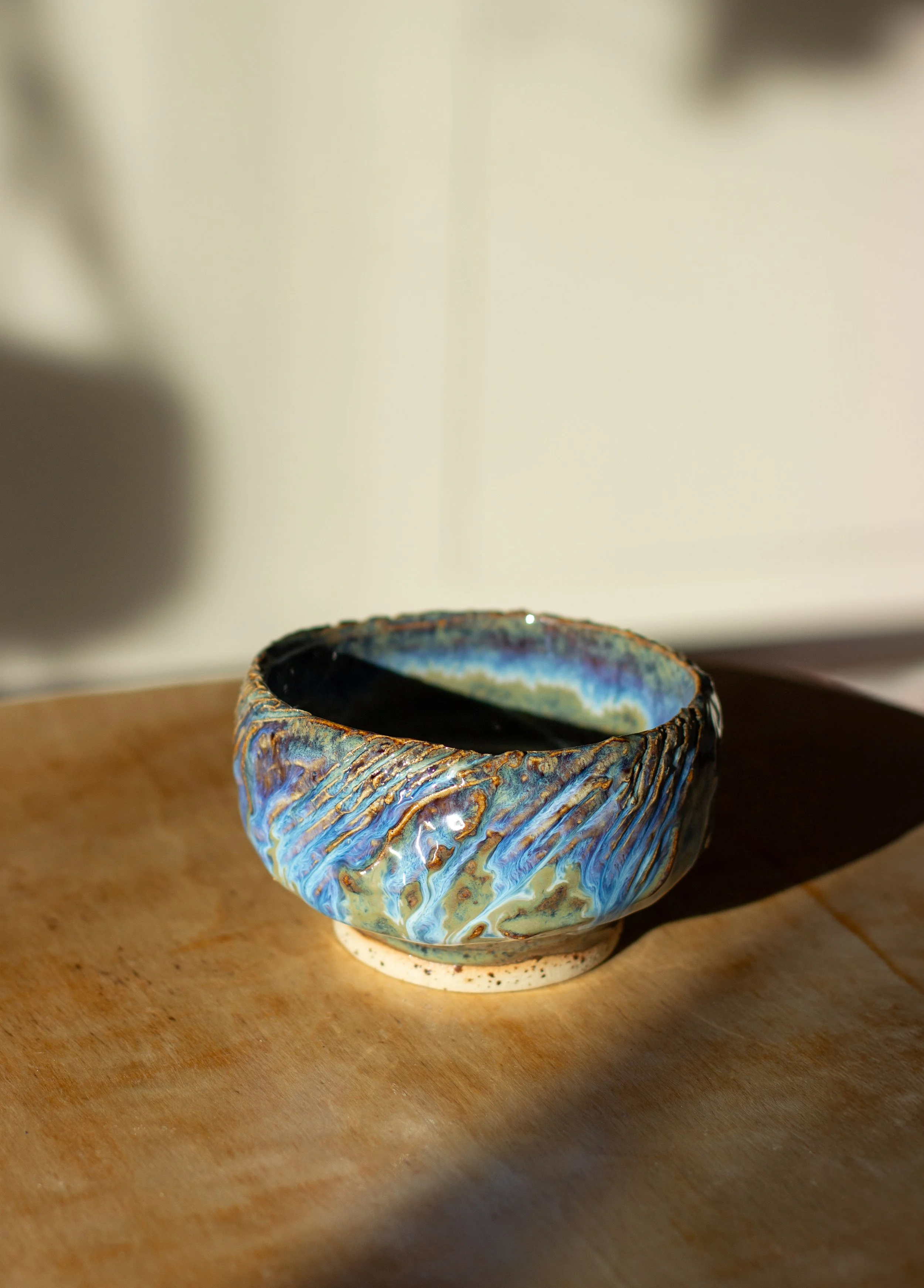 Käsintehty saviastia, jossa on sinisiä, vihertäviä ja ruskeita kuvioita, ja se on asetettu puiselle alustalle auringonvalossa.