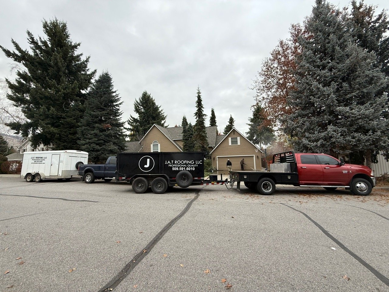 Line of vehicles including a trailer, a black trailer with J.A.T Roofing LLC branding, and a red flatbed truck, parked on a residential street with trees and houses in the background.