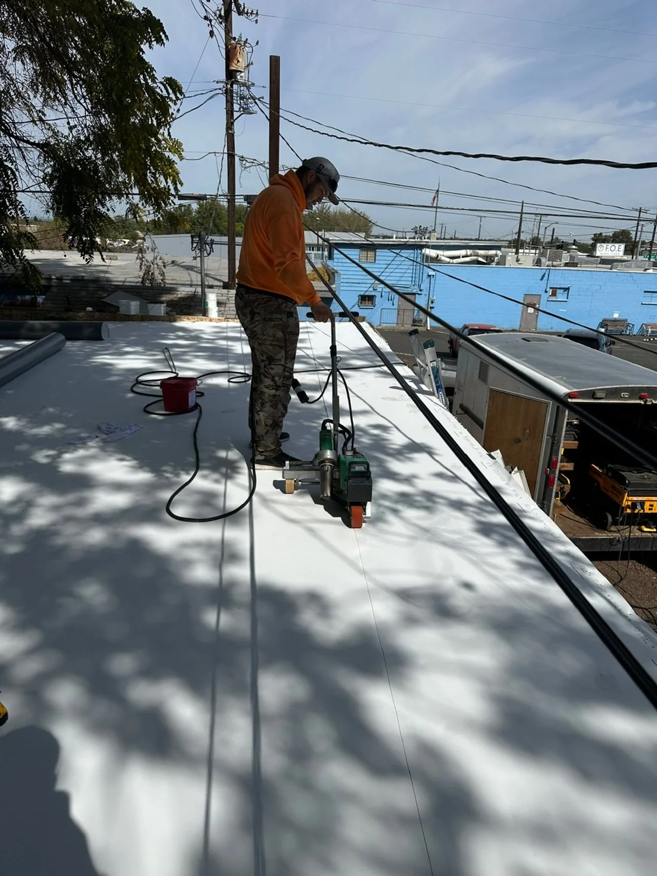 Man working on a flat roof with a power-cutting saw, surrounded by tools and construction equipment, in an industrial area with power lines and buildings in the background.