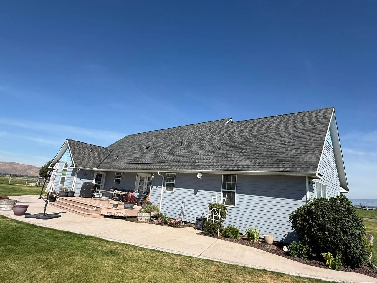 A blue house with a gray shingled roof, located in a grassy area with a concrete patio and outdoor furniture, under a clear blue sky.
