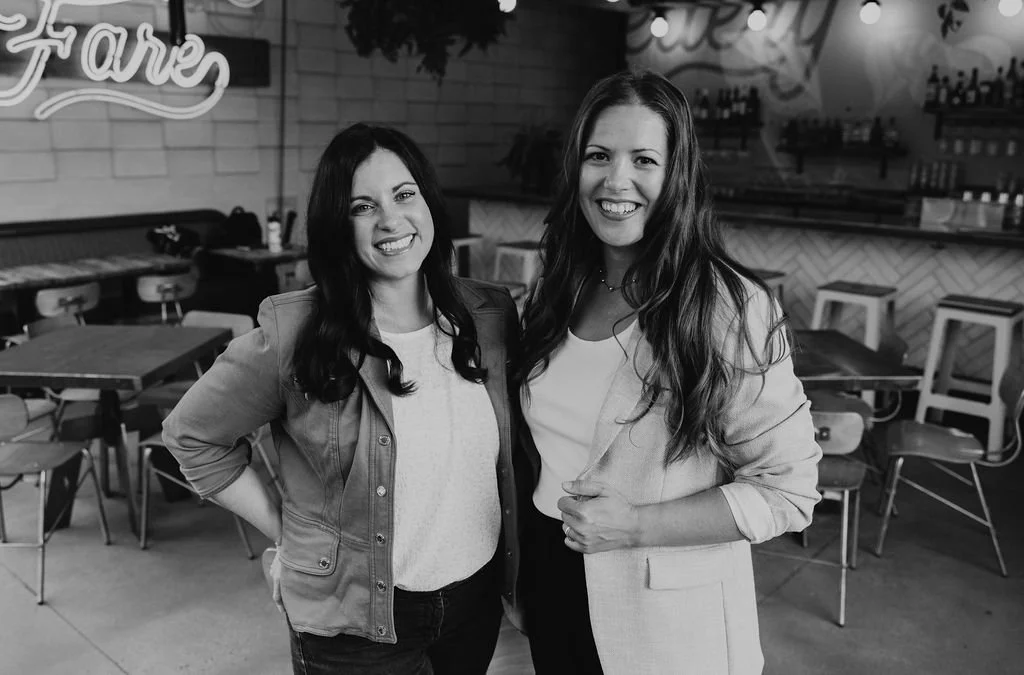 The cofounders of the Collective for Designers, Keri and Erin pose. They are two smiling women standing inside a cafe or restaurant, with tables and chairs in the background, in black and white.