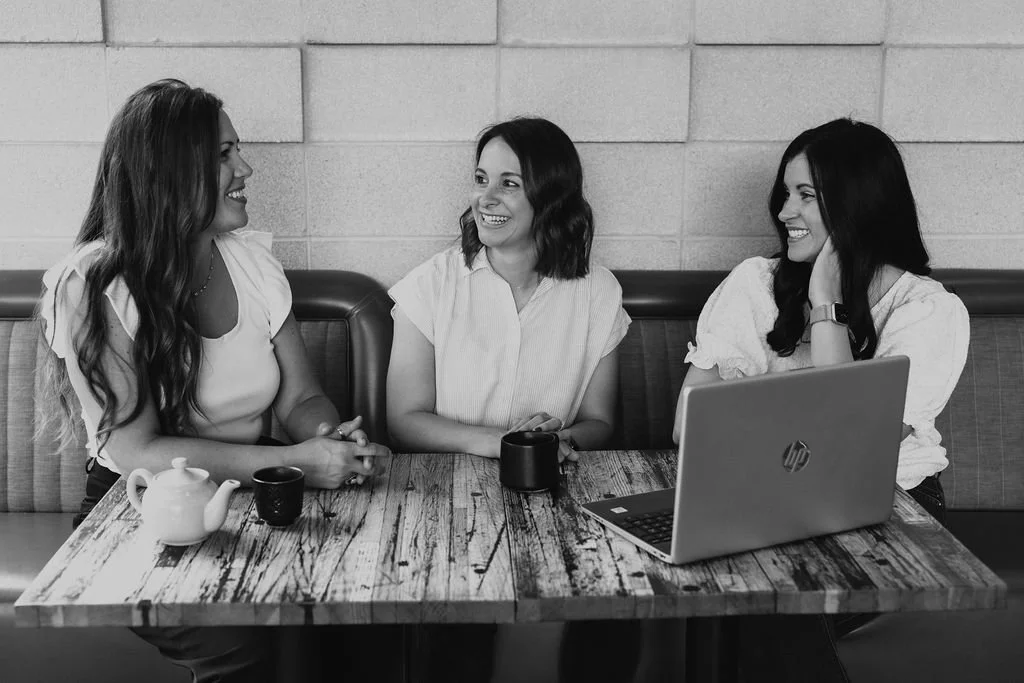 the collective for designers during a business consulting session with an interior designer. Three women sitting at a table, smiling and talking, with a laptop and coffee cups in front of them, in a casual setting.