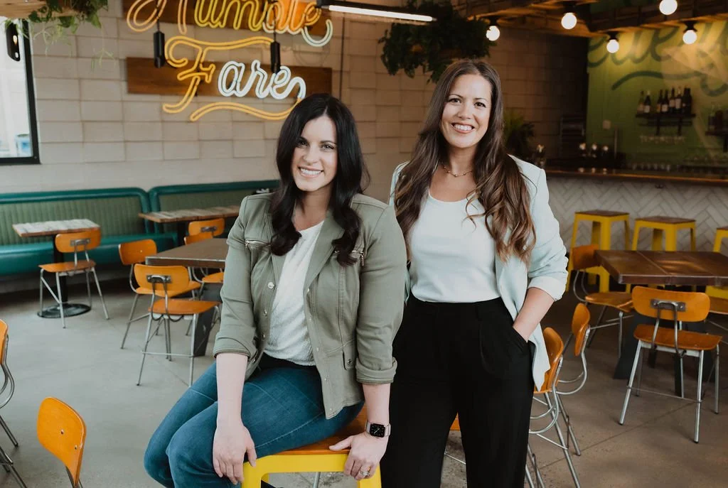 The Collective for Designers CoFounders (Keri Hinterreiter sitting, Erin Hildmann standing) smile at the viewer