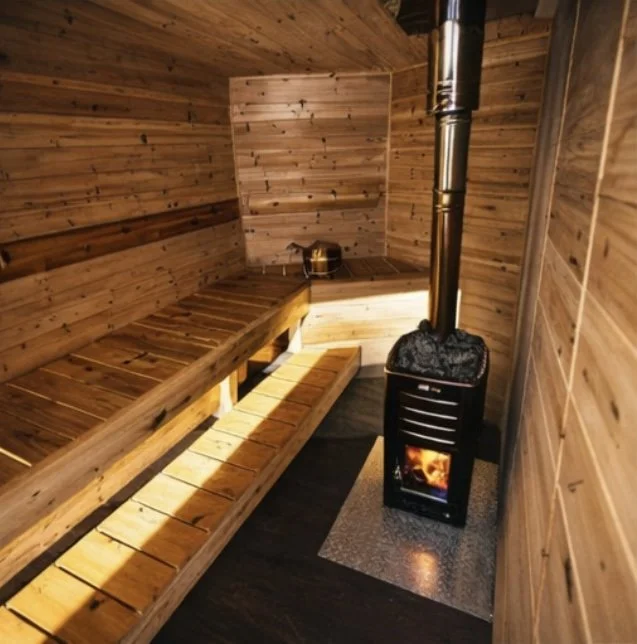 A small wooden sauna room with a bench along the left wall, a small window at the far end, and a wood-burning stove with a chimney pipe on the right, with burning logs visible inside.