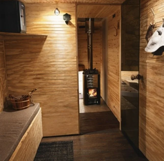 Wood-paneled interior with a small wood stove burning fire, a bench with a basket on it, and wall-mounted hooks with hats, all inside a cozy cabin.