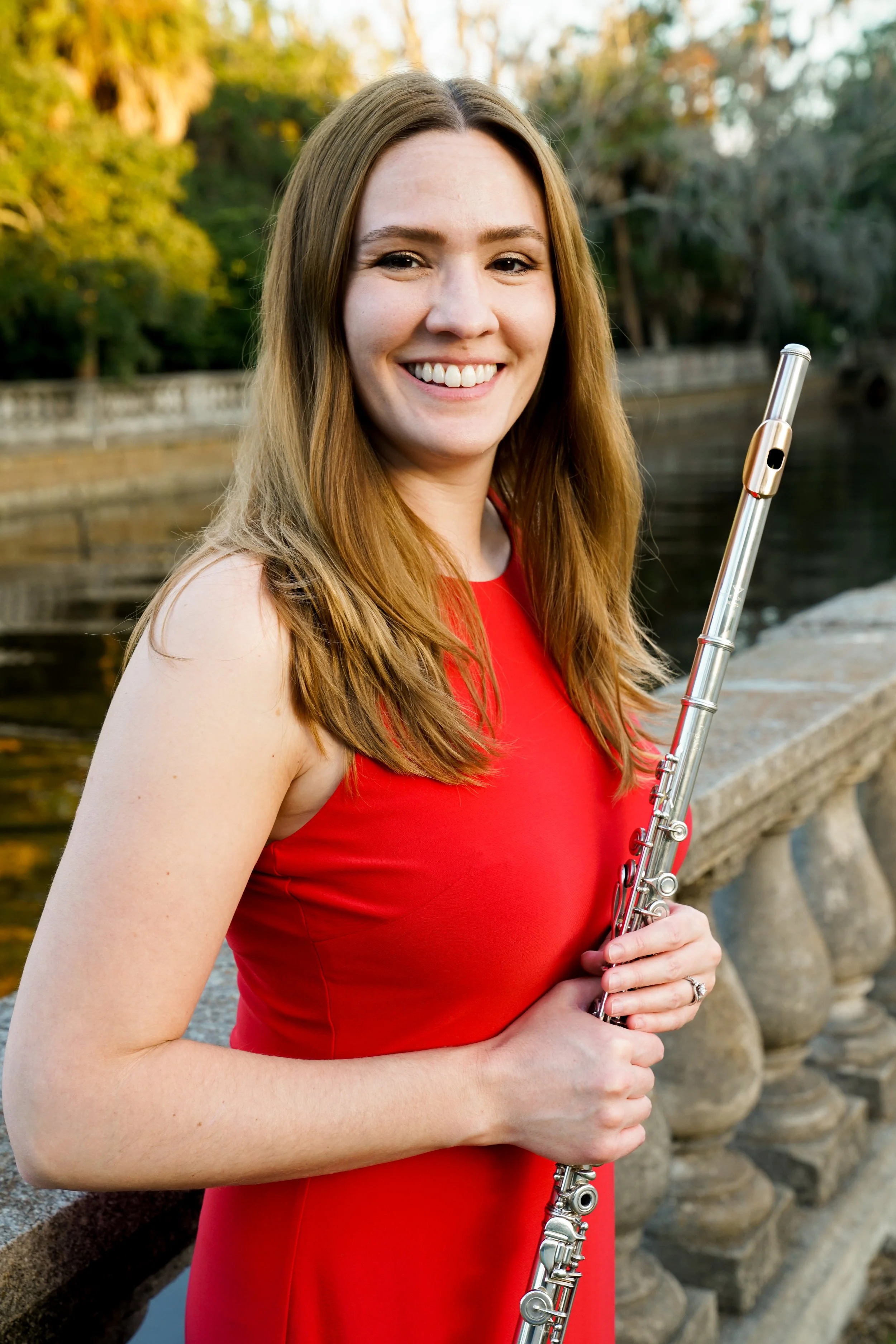 A young woman with long brown hair wearing a red sleeveless dress, holding a flute, standing outdoors near a stone railing by a body of water with trees in the background.