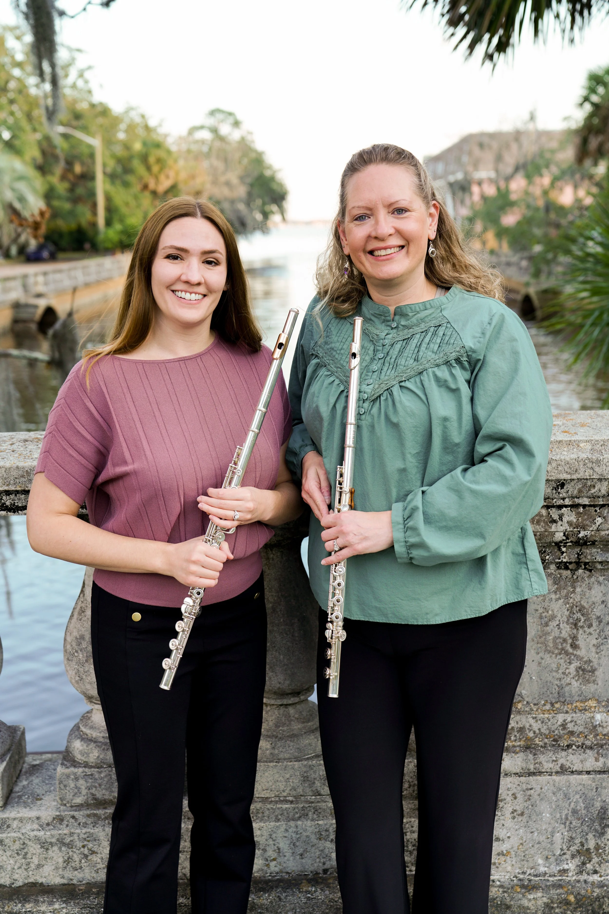 Two women standing outdoors on a stone bridge, holding flutes, smiling, with a river and trees in the background.