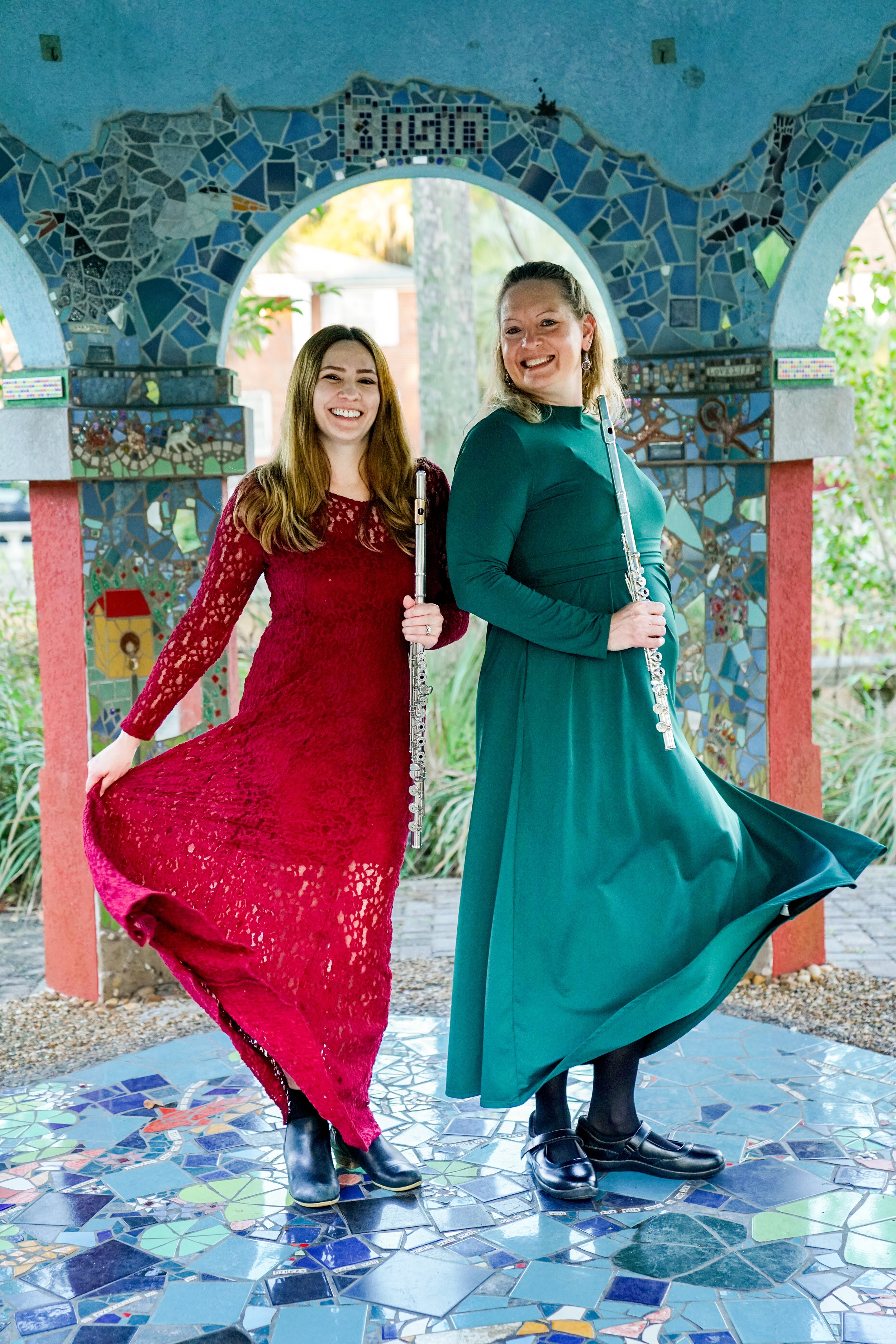 Two women in colorful dresses standing in front of a mosaic arch, each holding a flute, smiling at the camera.