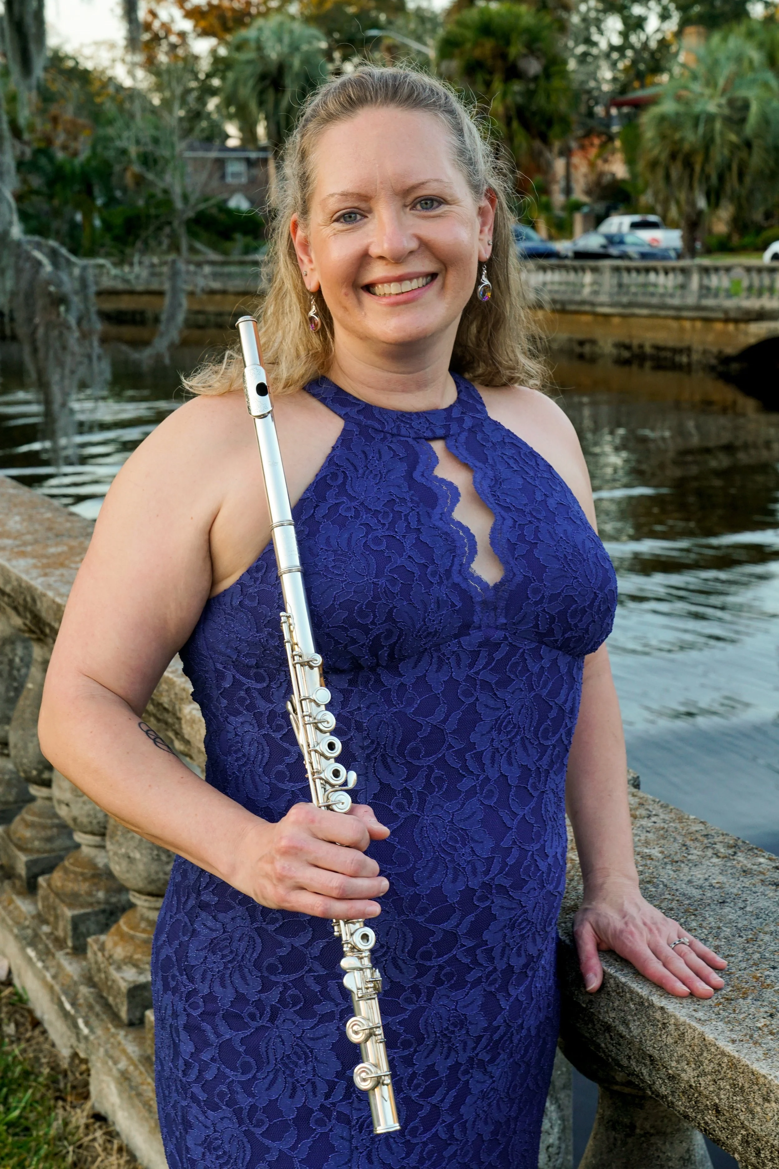 Woman in a blue lace dress holding a flute near a river with trees and cars in the background.