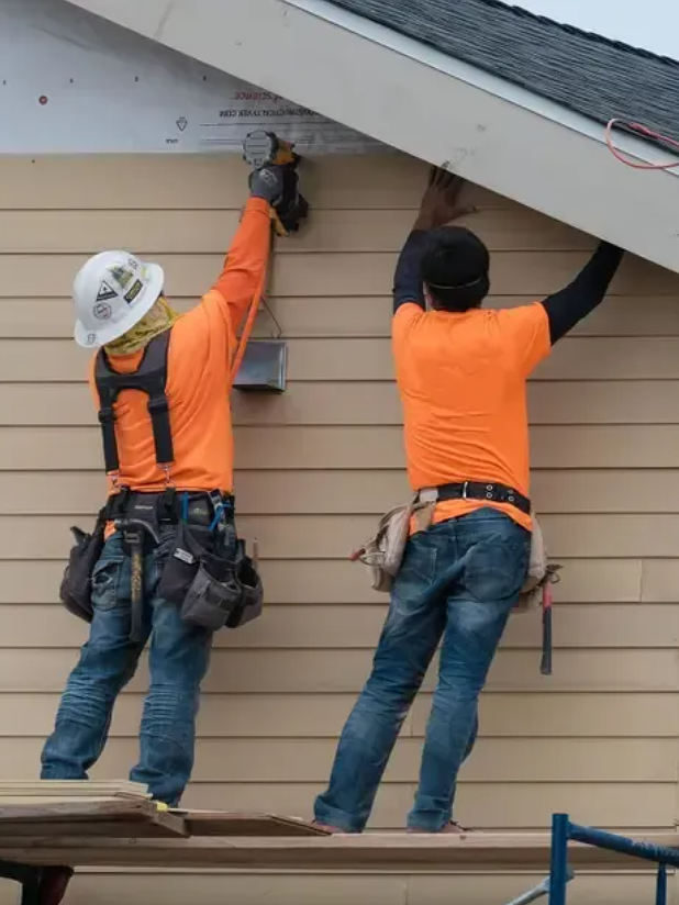 Two construction workers in orange shirts and tool belts, one with a white helmet, installing or repairing soffit beneath the roof of a building.