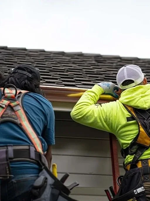 Two construction workers, one in a blue shirt and the other in a neon green hoodie, are working on the roof. They are installing or repairing shingles on the roof of a house, with tools and safety equipment visible.