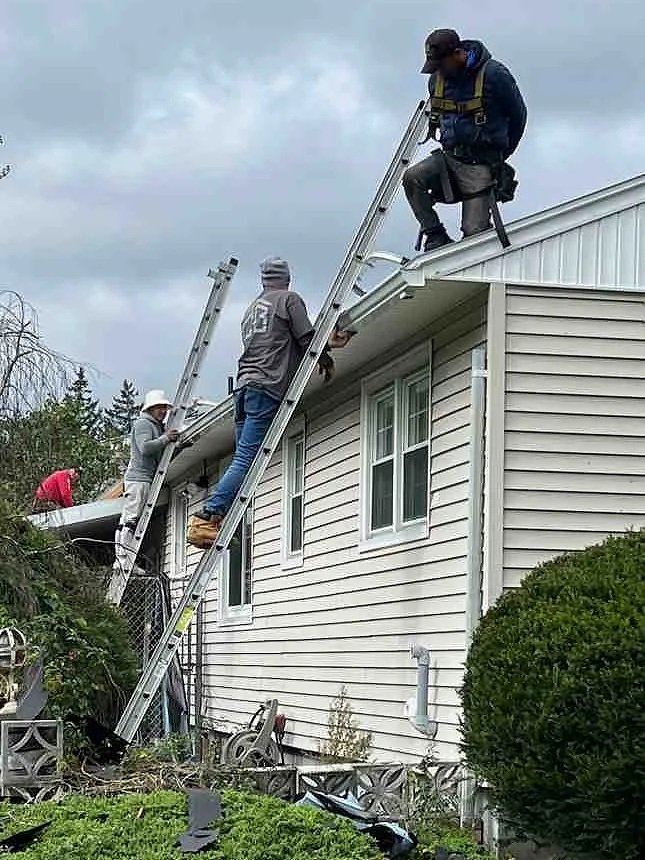 Construction workers on ladders repairing the roof of a house during overcast weather.