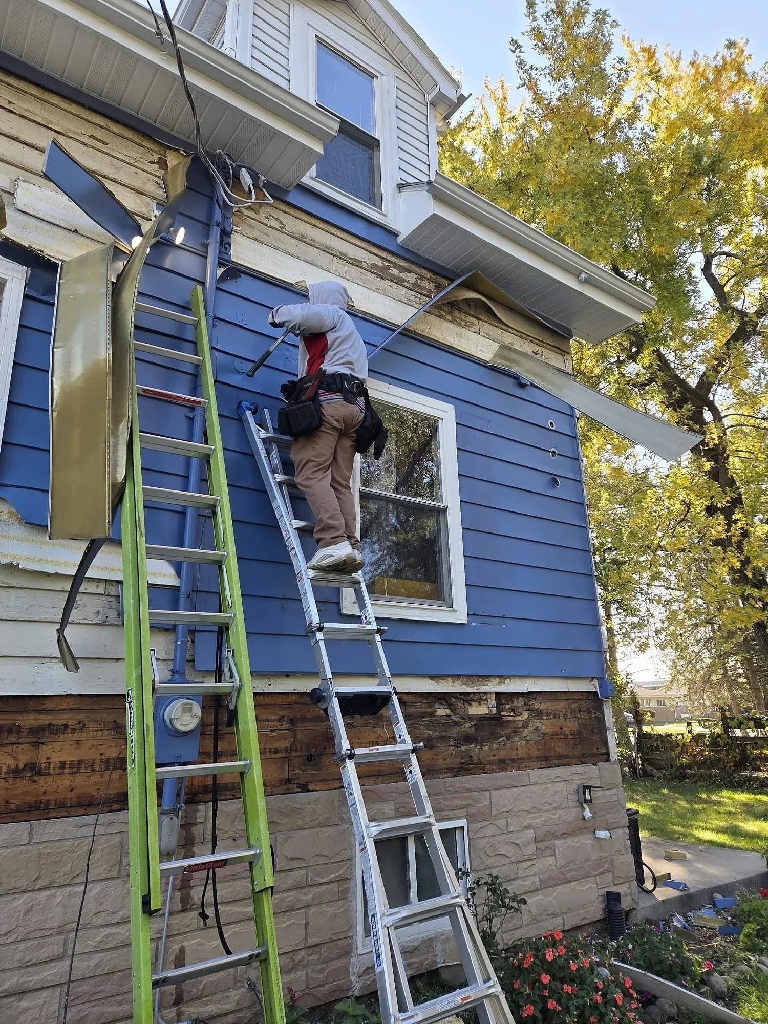 A worker installing or repairing siding on a two-story house, standing on an extension ladder, with two ladders leaning against the house and tools attached to his belt.