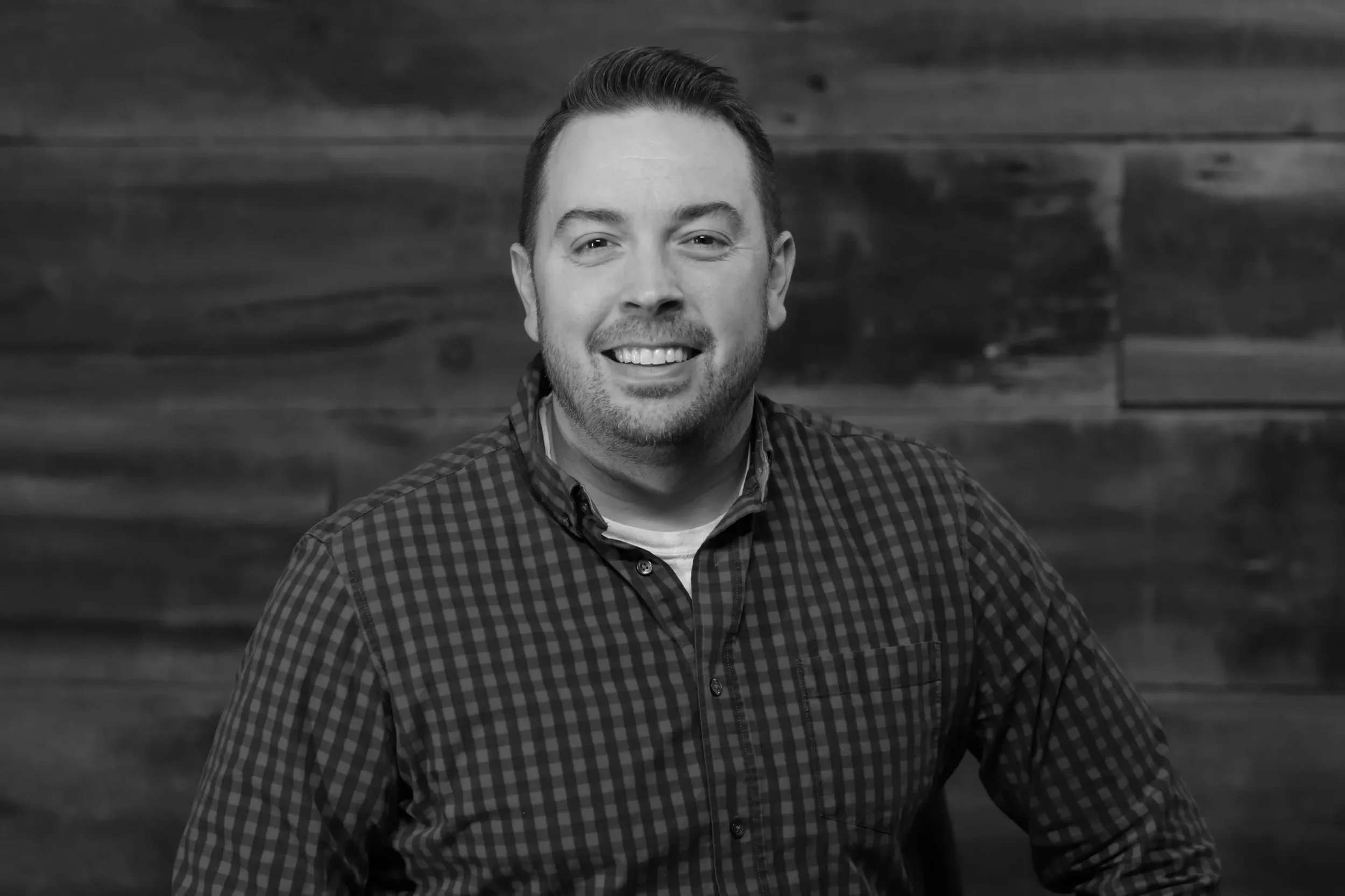 A smiling man with short hair and beard, wearing a checkered shirt over a T-shirt, standing in front of a wooden wall.