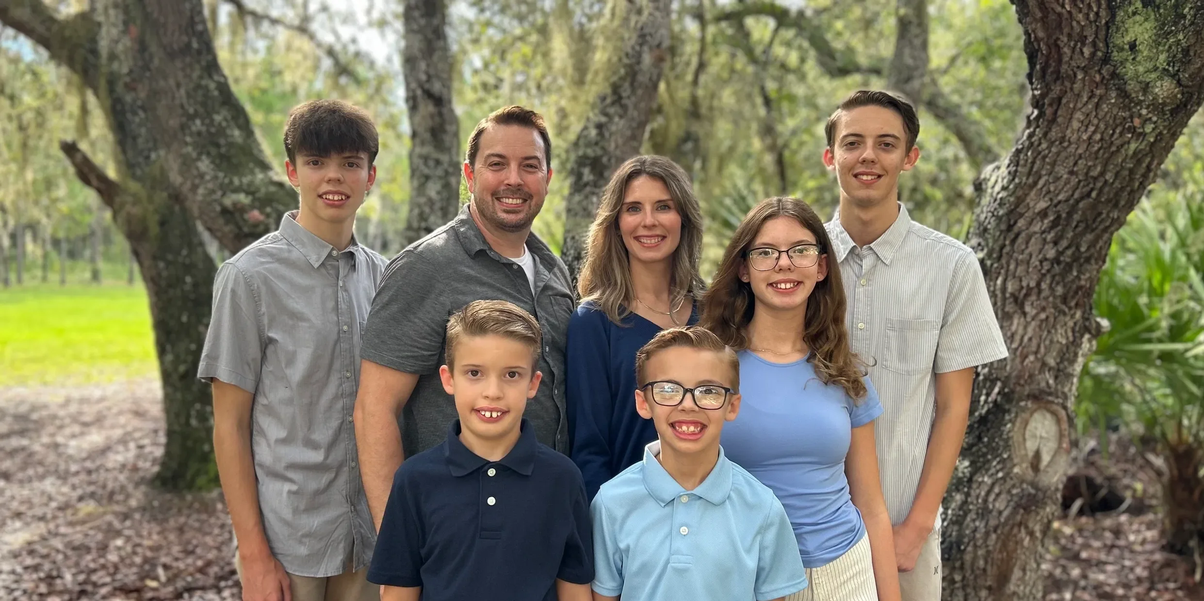 A family of seven posing outdoors in a wooded area with large trees and green foliage, smiling at the camera.
