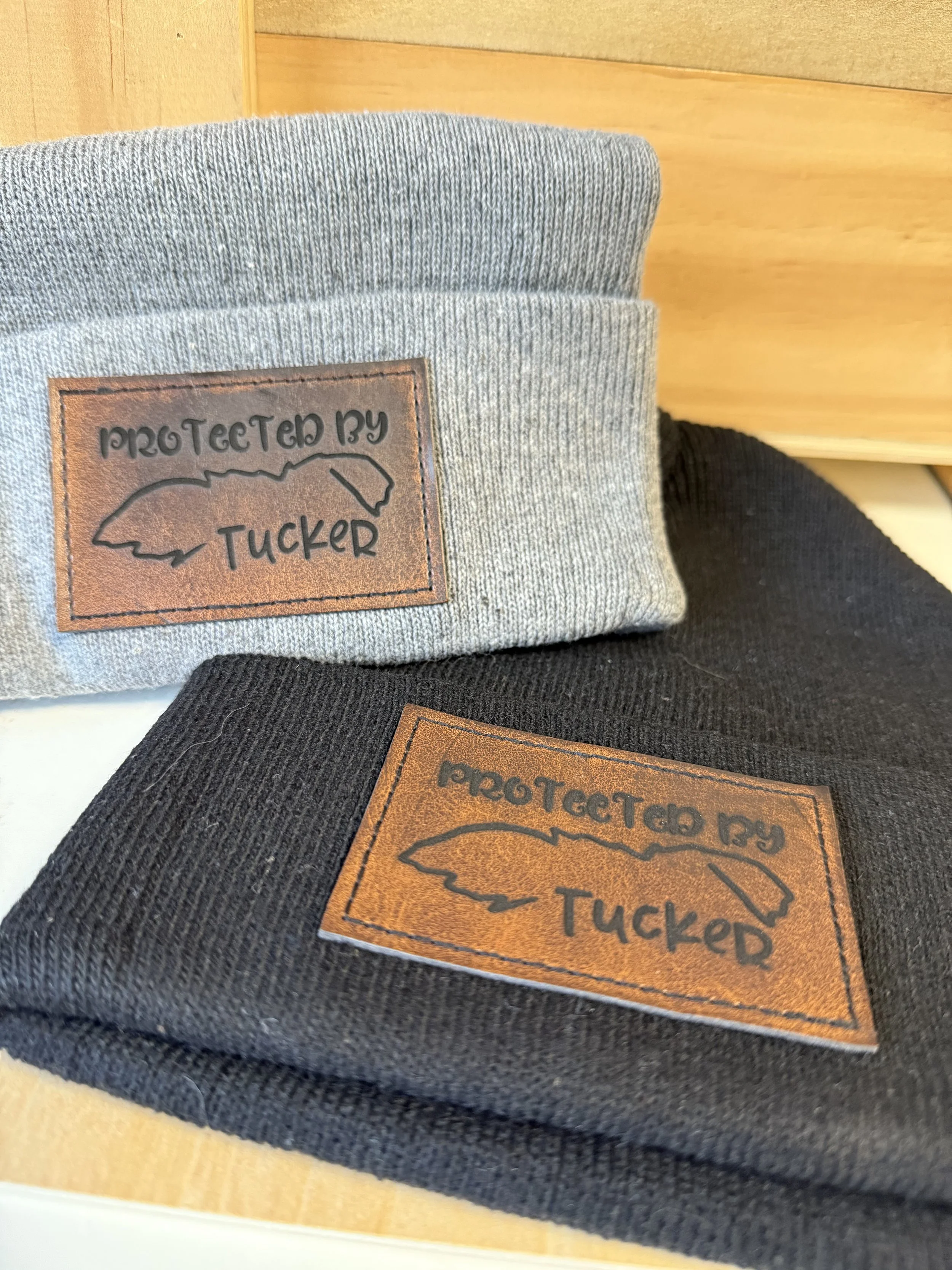 Two beanies- black and gray- with rustic brown, rectangle, laser engraved patches that say, "protected by tucker." Custom name and outline of the pet ears. 