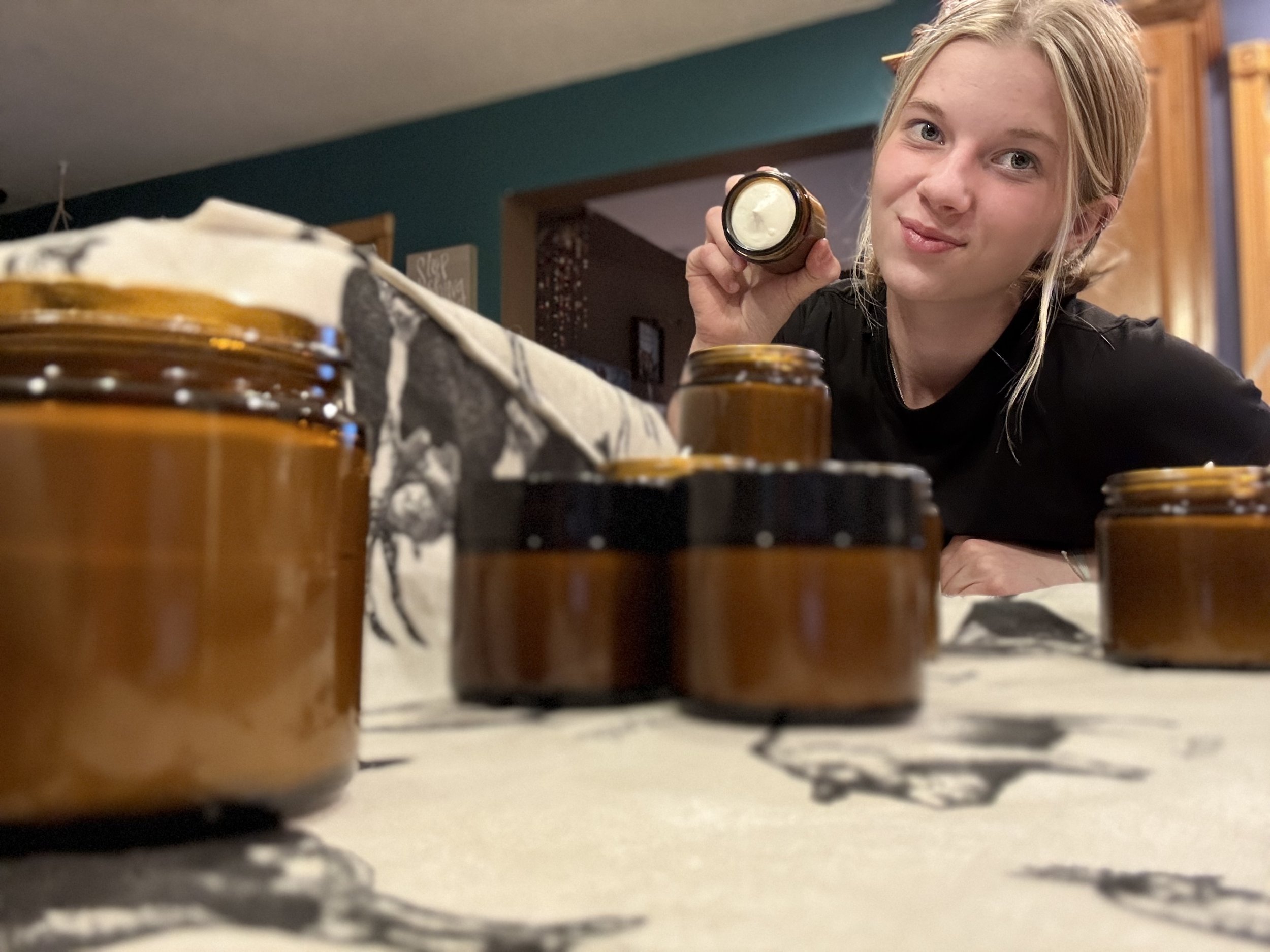 A young woman holds a small jar of whipped tallow while looking at the camera, surrounded by several similar jars on a table covered with a cloth.
