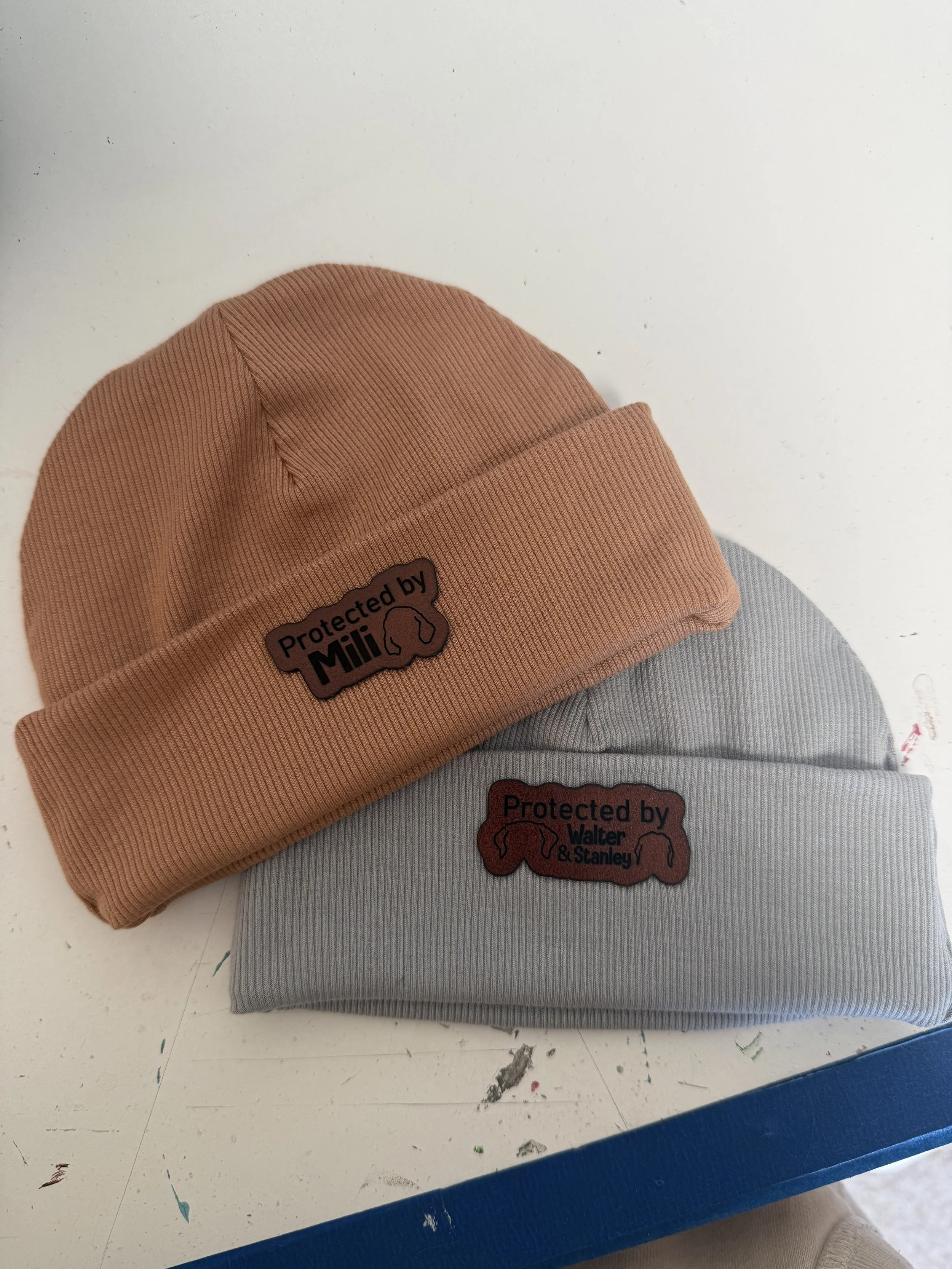 Orange and grey newborn hats with rustic brown, laser engraved patch. Patch includes custom dog ear outline and "protect by" custom pet name. 