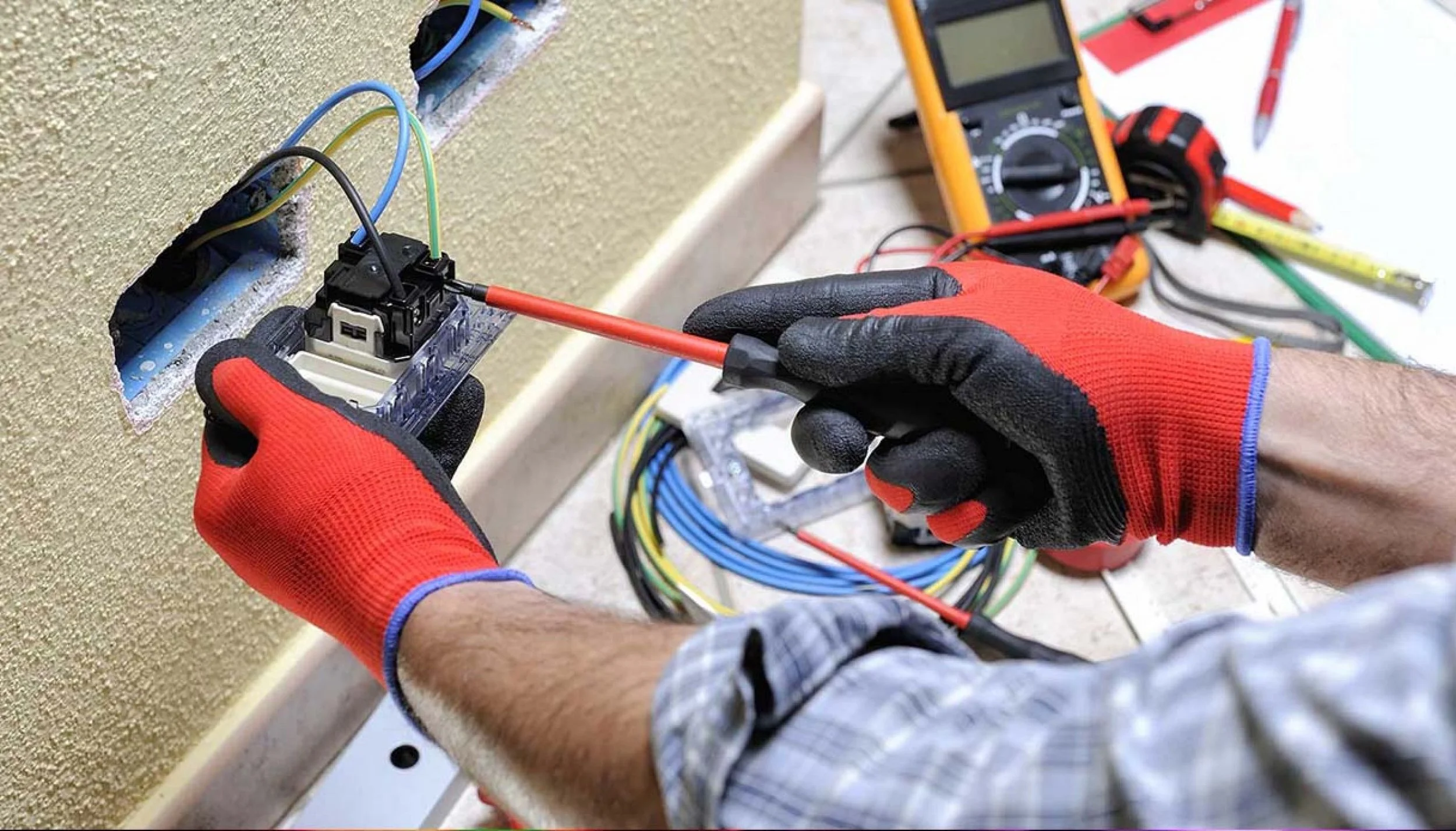 A person wearing black and red gloves is using a screwdriver to work on an electrical outlet box with wires inside. There are electrical tools and a multimeter on the floor nearby.