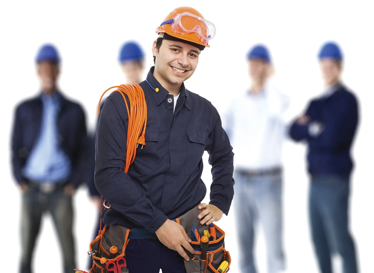 A smiling male worker wearing a safety helmet, goggles, and a tool belt, with a group of blurred business professionals in the background.