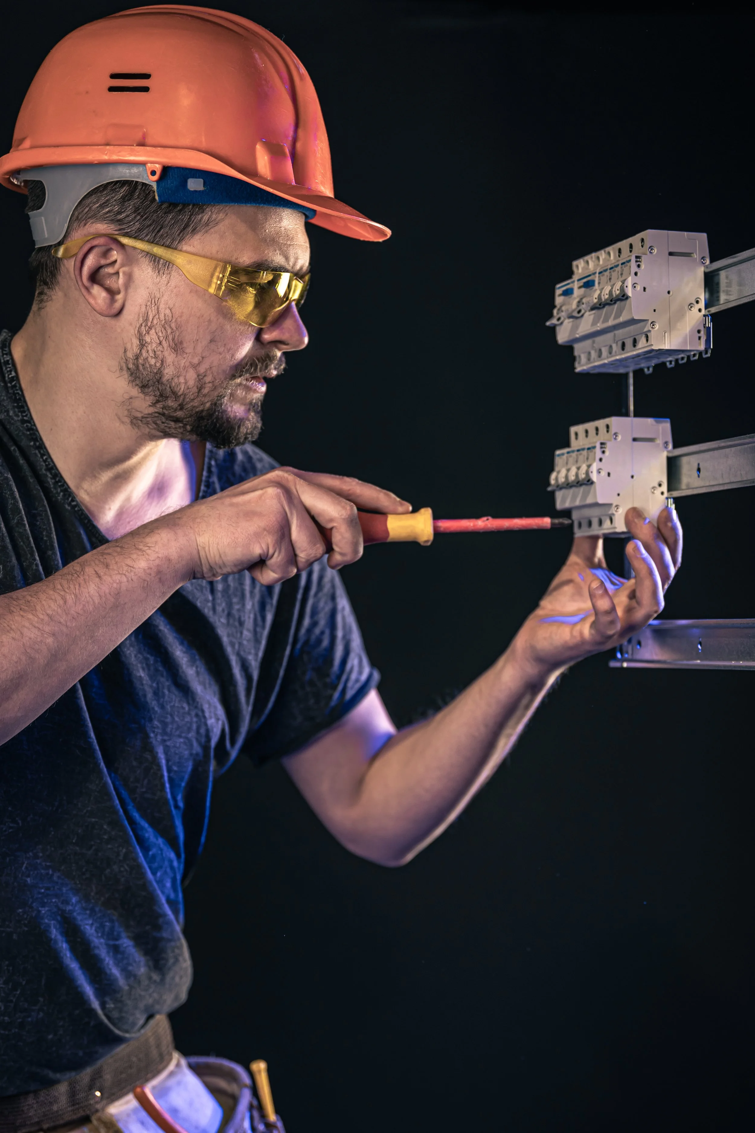 A construction worker wearing a red hard hat, yellow safety glasses, and a black shirt, using a screwdriver on metal electrical components against a dark background.