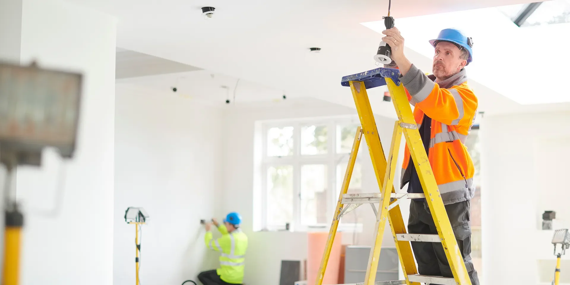 Construction worker on a yellow ladder installing or fixing a light fixture in a white room.