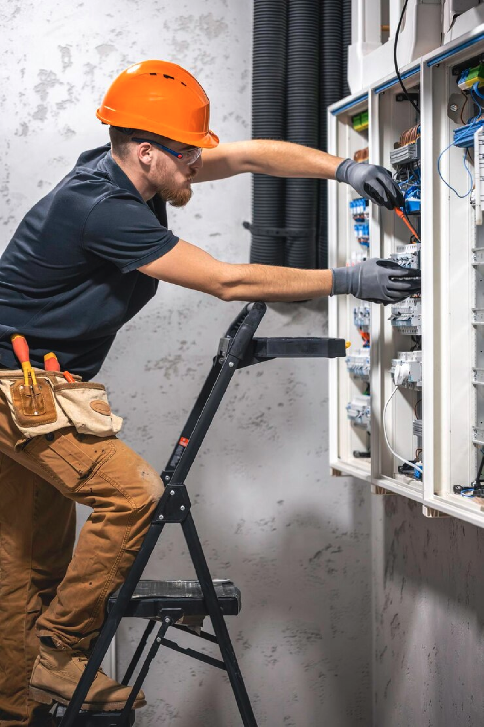 A male electrician wearing an orange safety helmet and gloves working on an electrical panel with various wires.