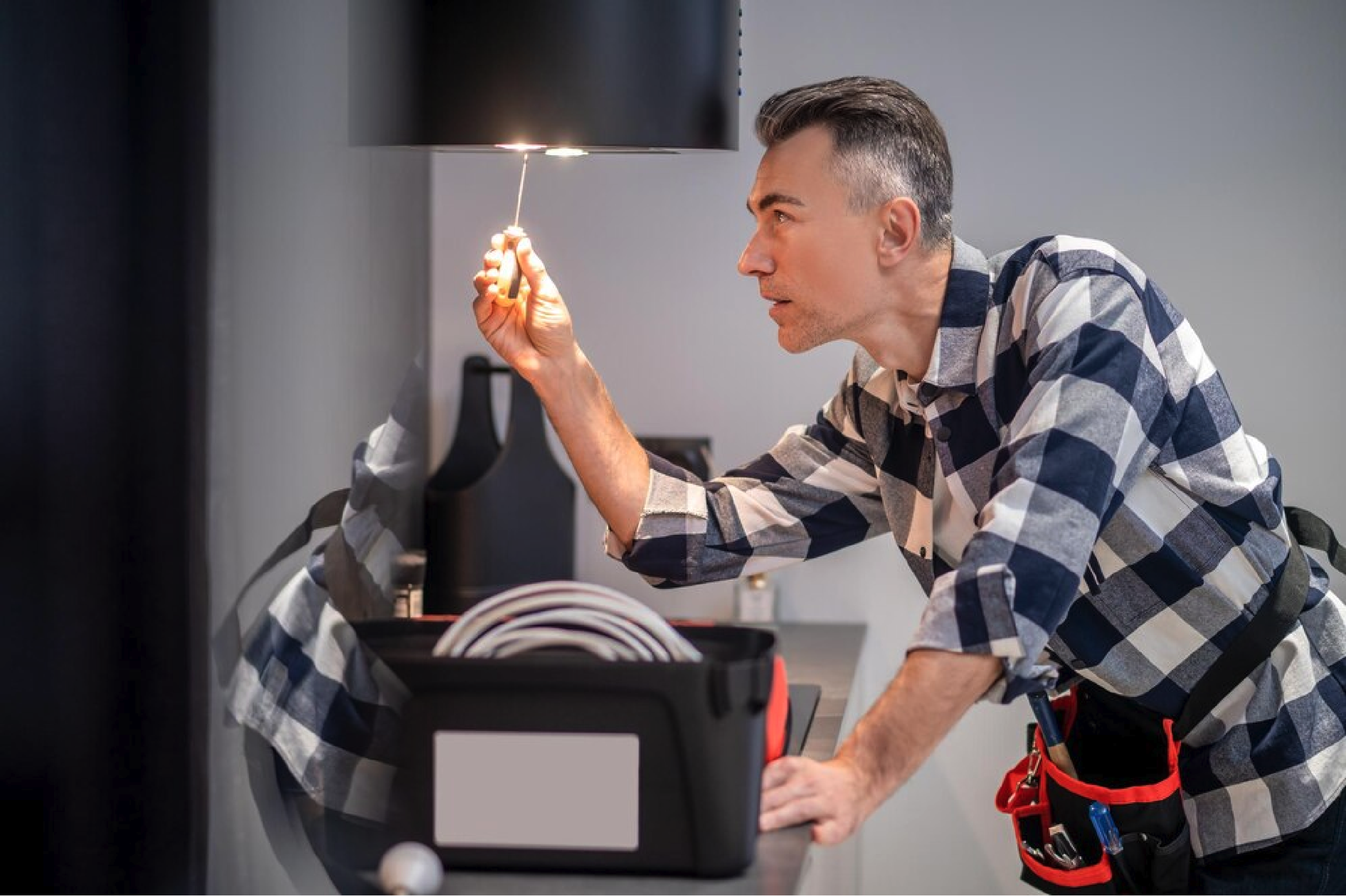 A man in a plaid shirt inspecting a ceiling light fixture with a flashlight in a modern room, with a toolbox on the table.