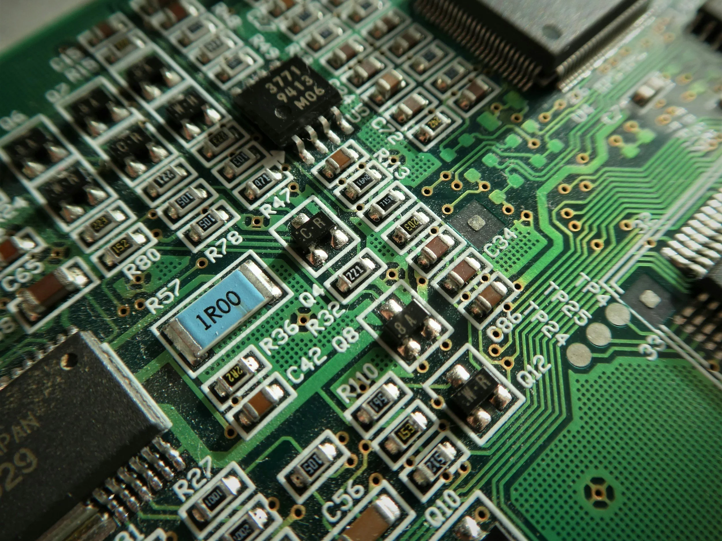 Close-up of a green circuit board with various resistors, capacitors, and integrated circuits, including a blue resistor marked '1R00'.