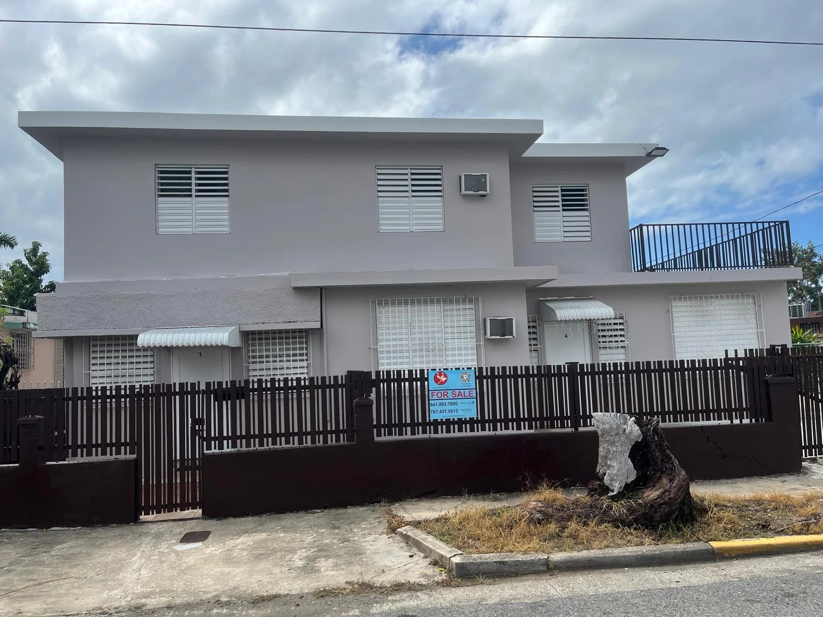 A two-story white house with metal window shutters, air conditioning units, and a fenced yard. For sale sign on the fence, cloudy sky, and a large tree stump in front.