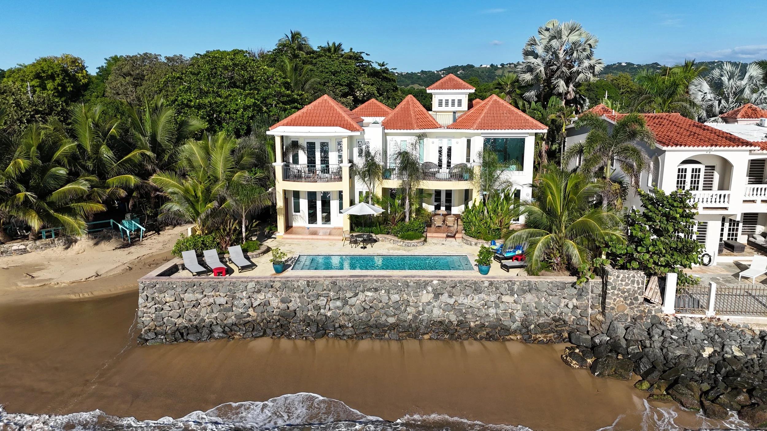 Yellow house with red tile roof, surrounded by palm trees and green foliage, with a black gate in a sunny setting.