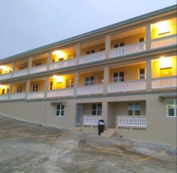 A multi-story motel or apartment building with three floors, lit by yellow lights, with a small parking lot in front and a trash can near the entrance.