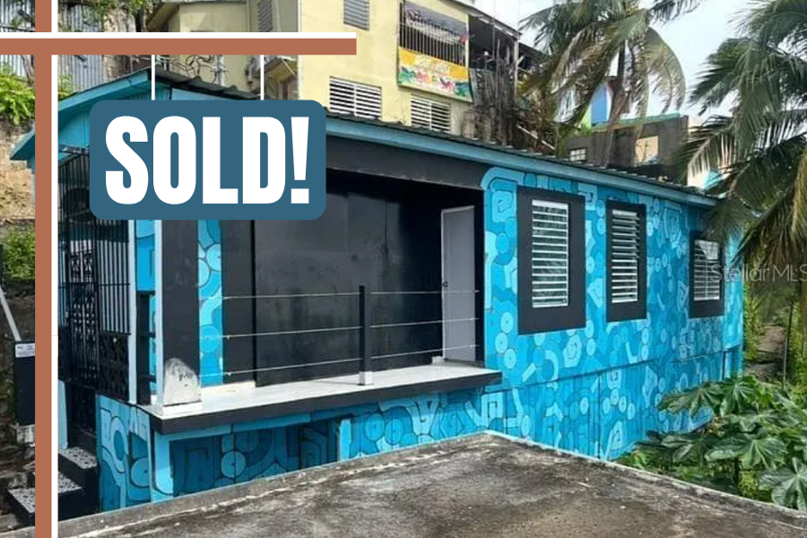 Colorful house with a blue patterned exterior, black window shutters, and an outdoor balcony with black railing, surrounded by palm trees.