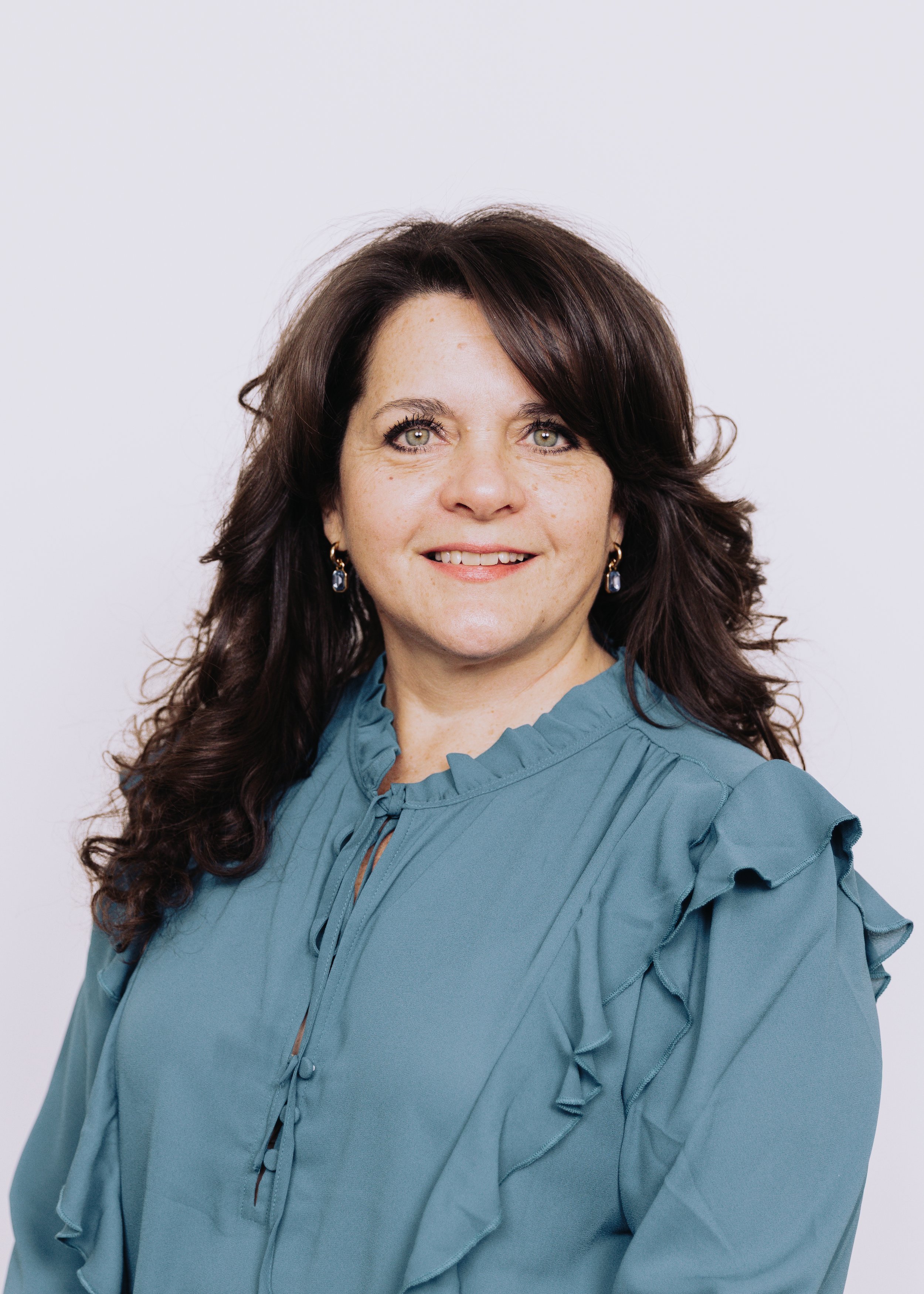 A woman with long dark hair wearing a blue ruffled blouse and earrings, smiling against a plain white background.