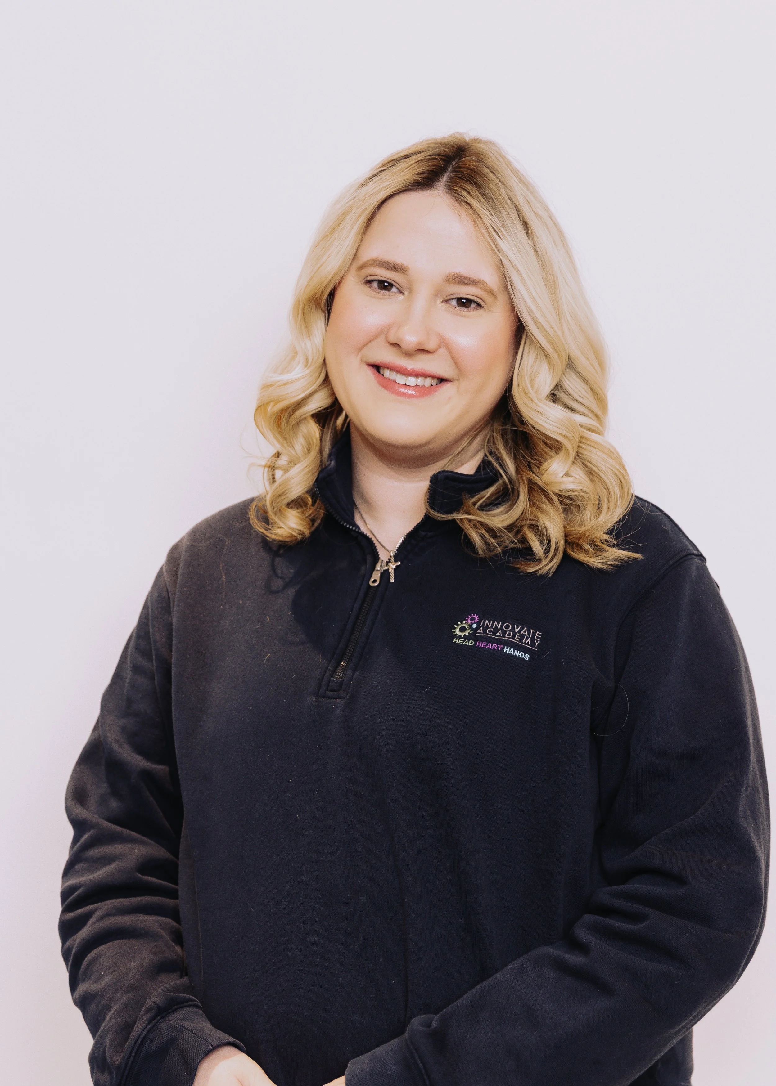 A woman with blonde hair smiling, wearing a black zip-up jacket with the 'Innovate Academy' logo.