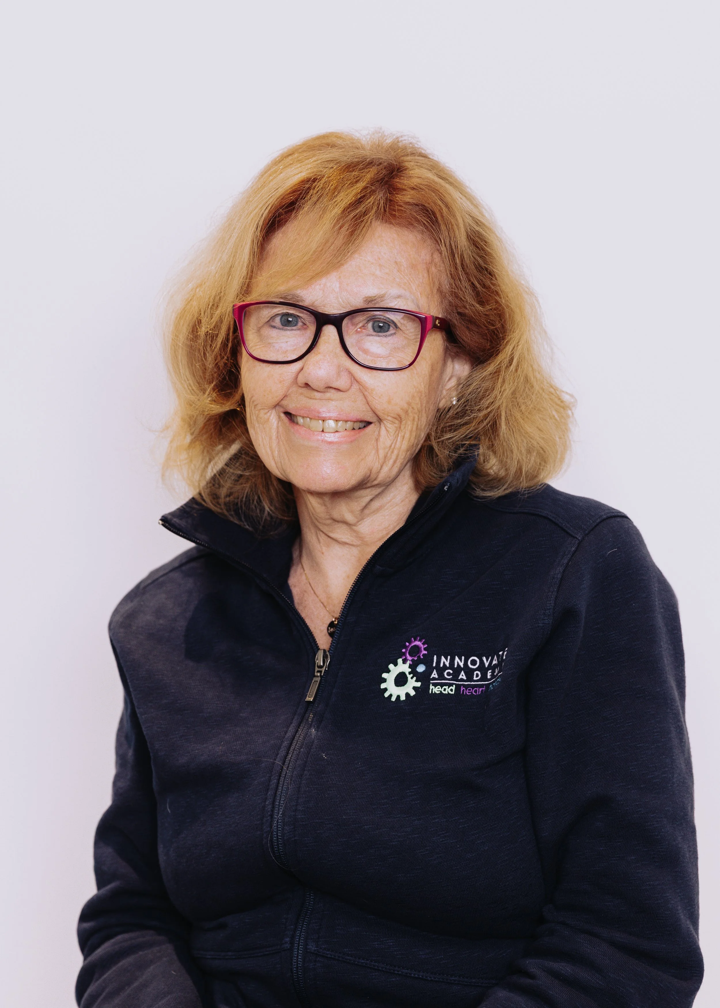 A smiling woman with red hair, wearing glasses and a black jacket with a logo that reads 'Innovative Academy' and 'head hear'.