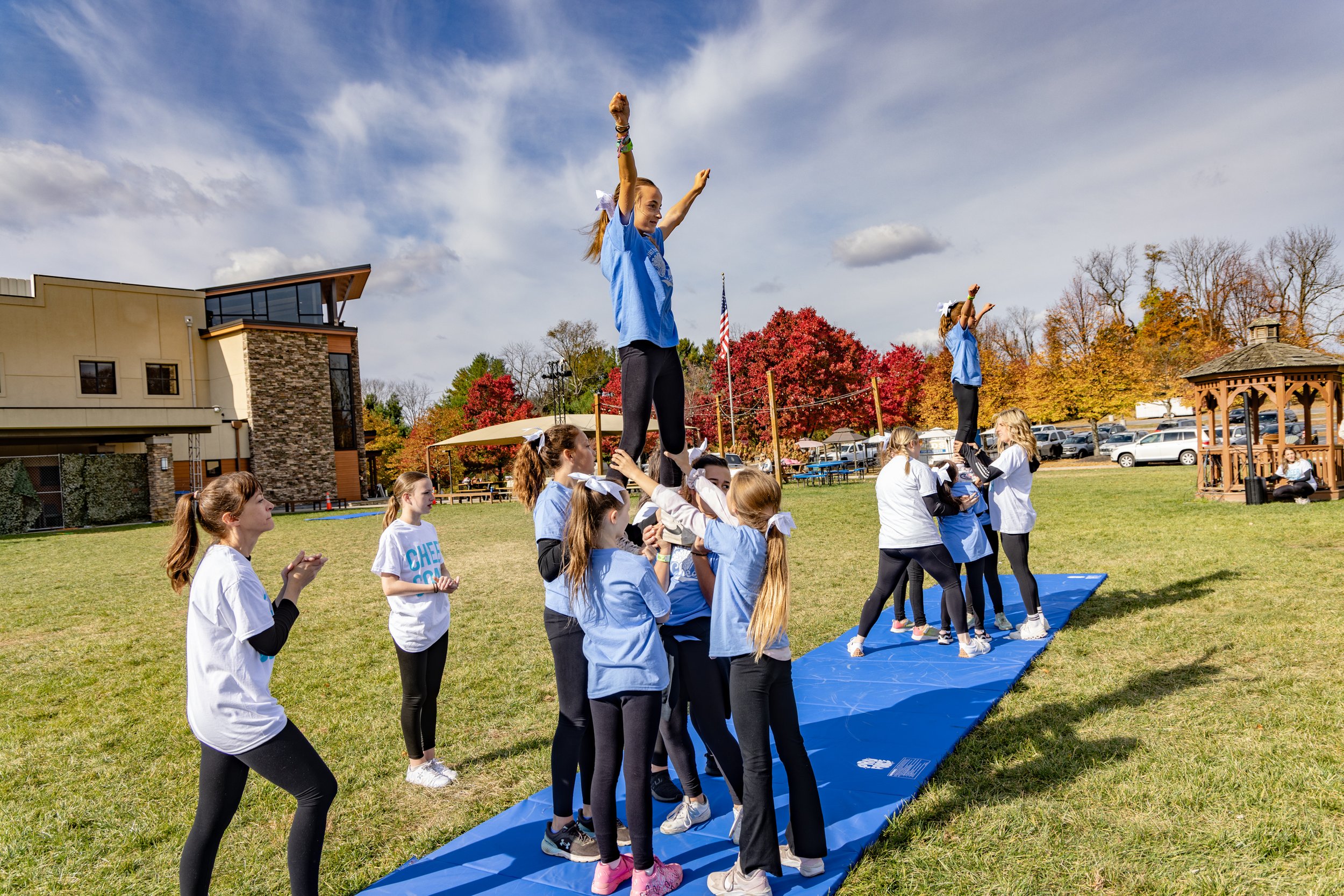 Cheerleading team practicing stunts outdoors on a blue mat with autumn trees and a building in the background.