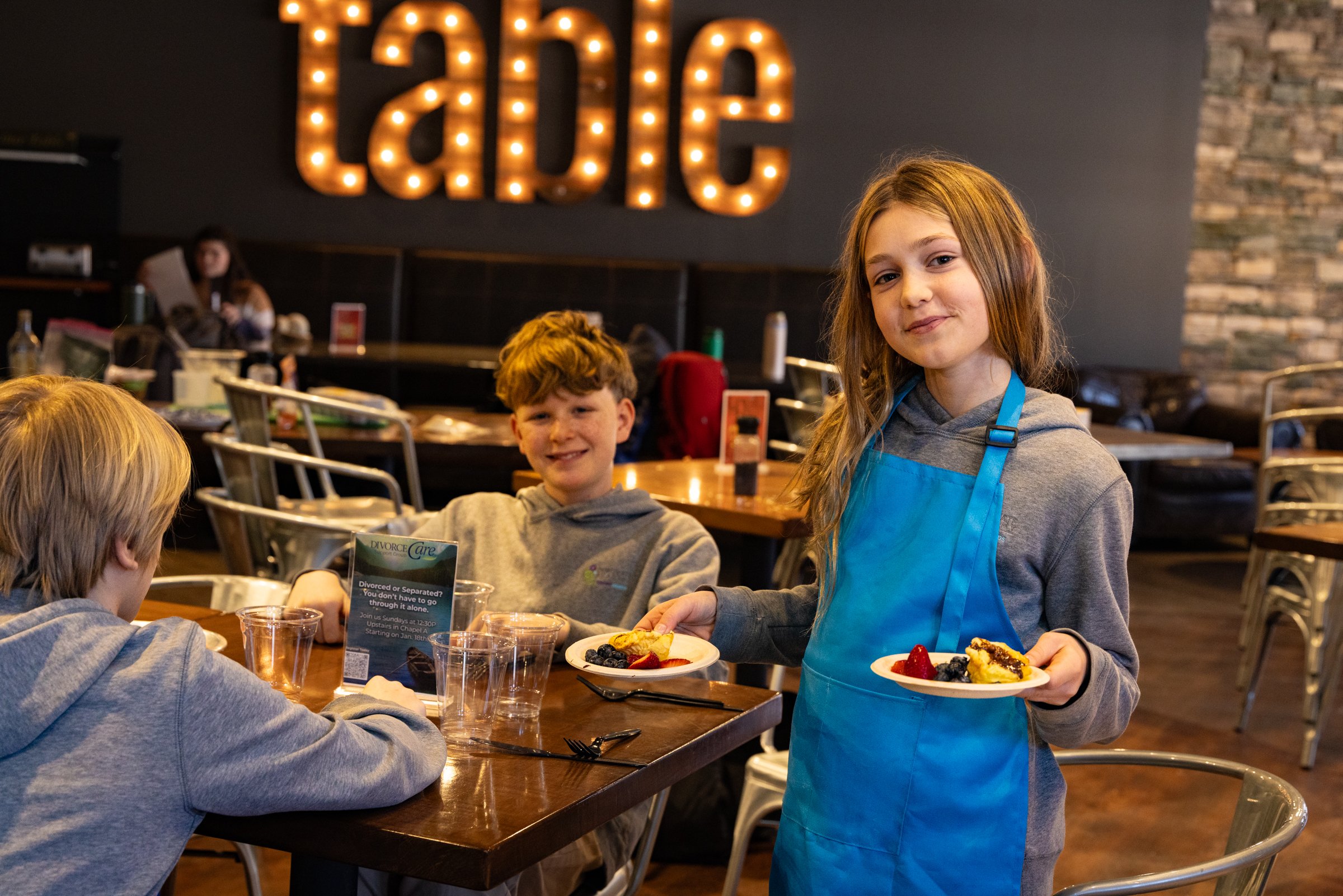 A young girl wearing a blue apron and smiling while holding two plates of dessert café in a restaurant with three other children at a table, one girl and two boys, with a large sign glowing with the word 'taste' in the background.