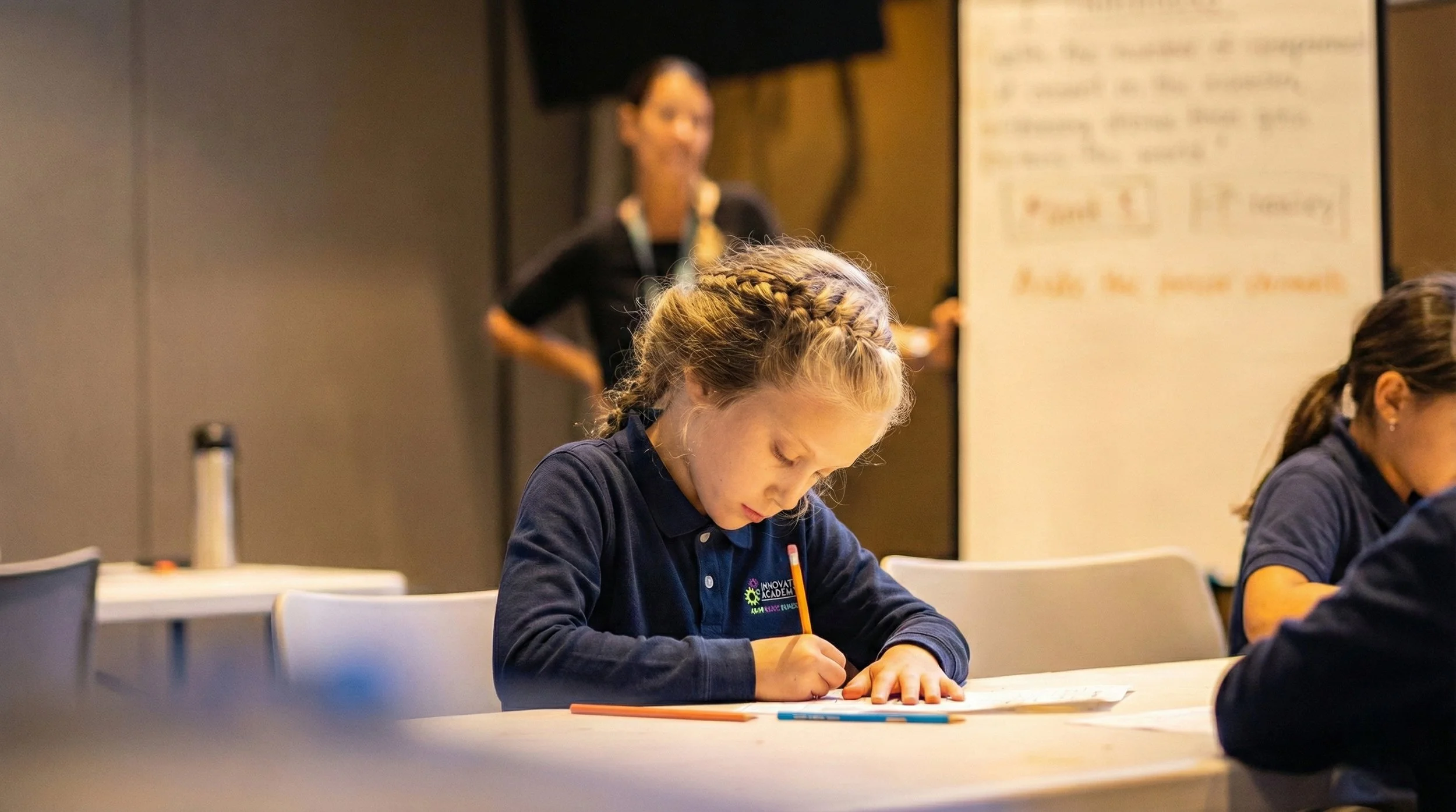 Young girl with braided hair writing on paper at a table in a classroom setting, with a woman standing in the background near a whiteboard.