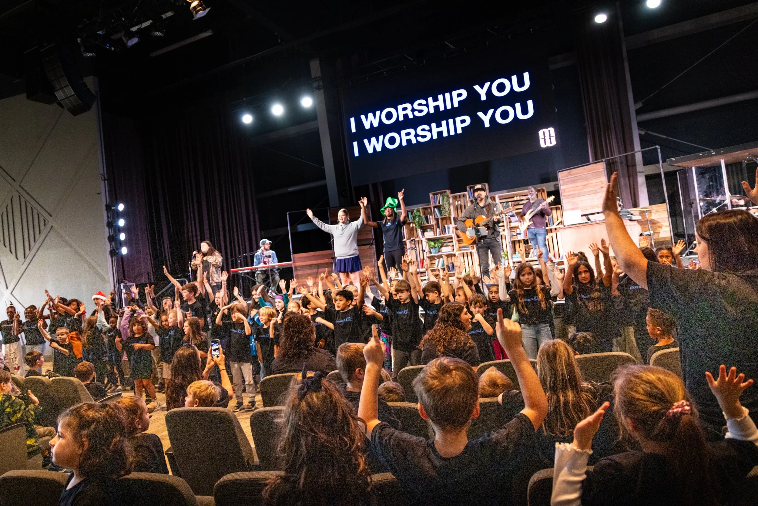 Children and adults participate in a church service or concert with raised hands, some on stage singing and playing music, and a large screen displaying the words "I worship you".