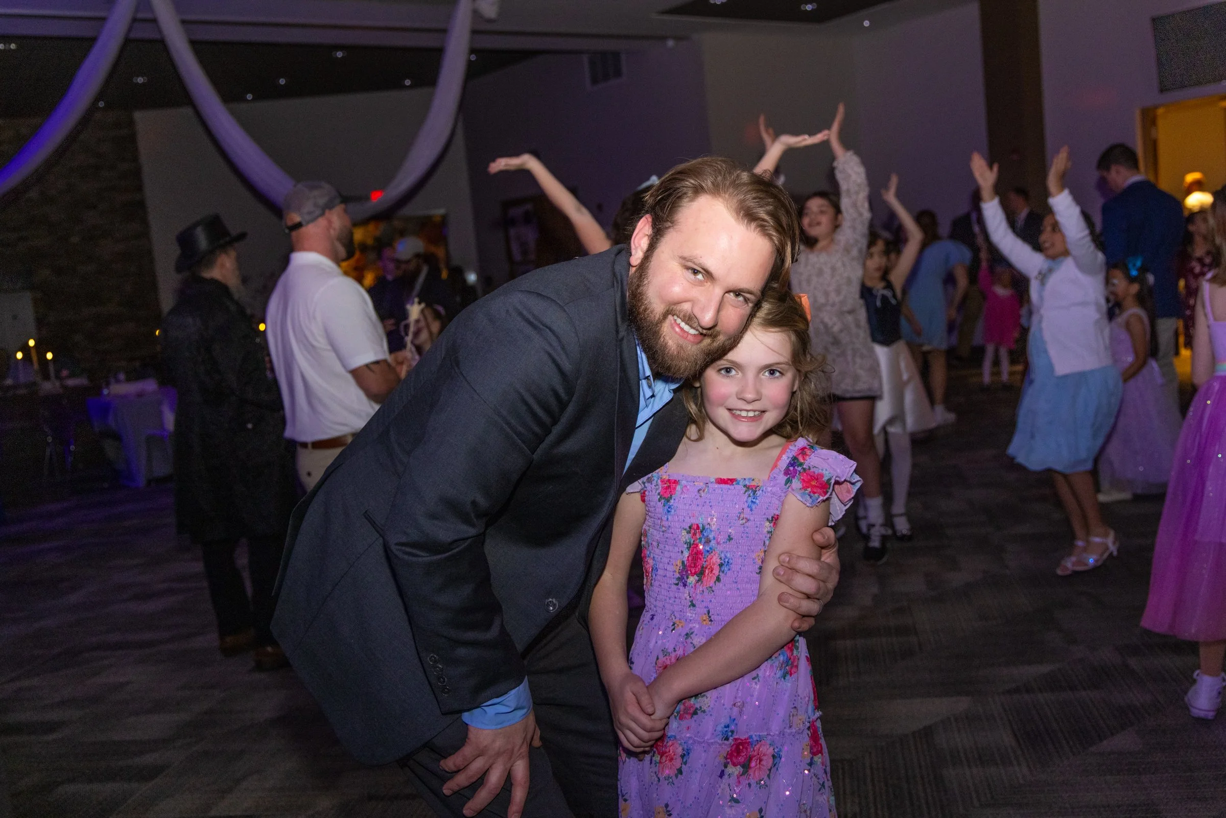 A man in a dark suit and a young girl in a floral dress smiling and dancing at a party or celebration.