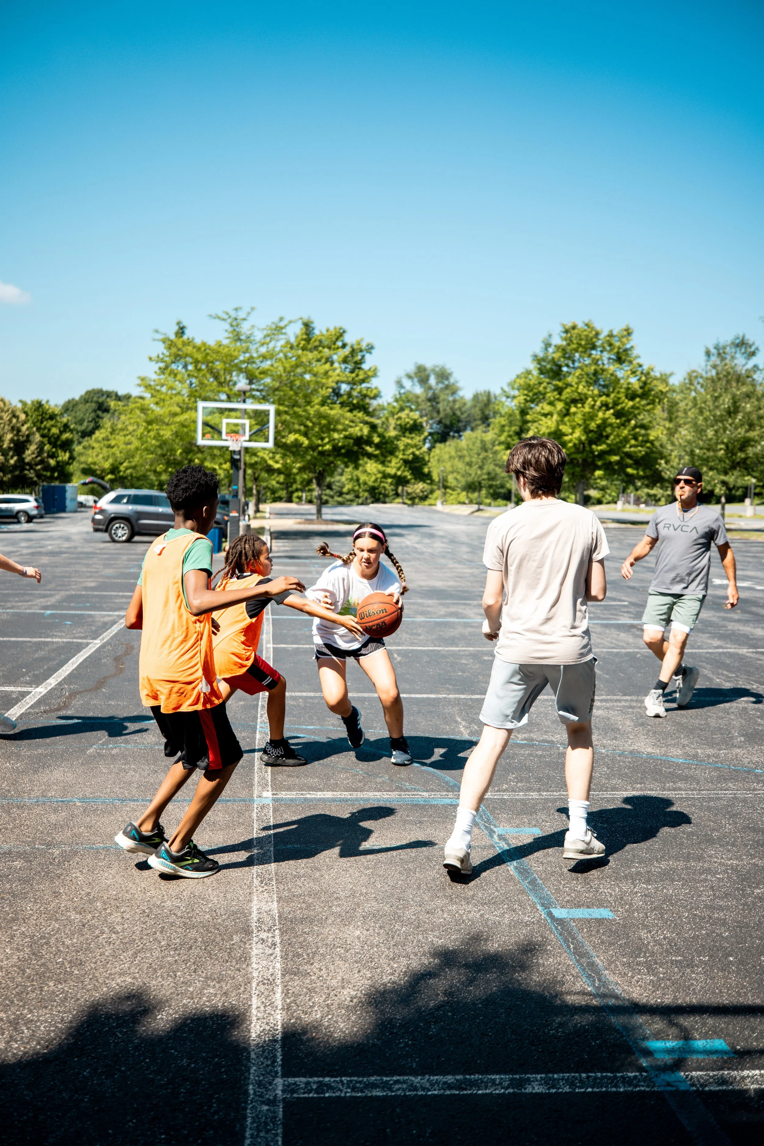 Kids playing basketball on outdoor court with trees and cars in the background under clear blue sky.