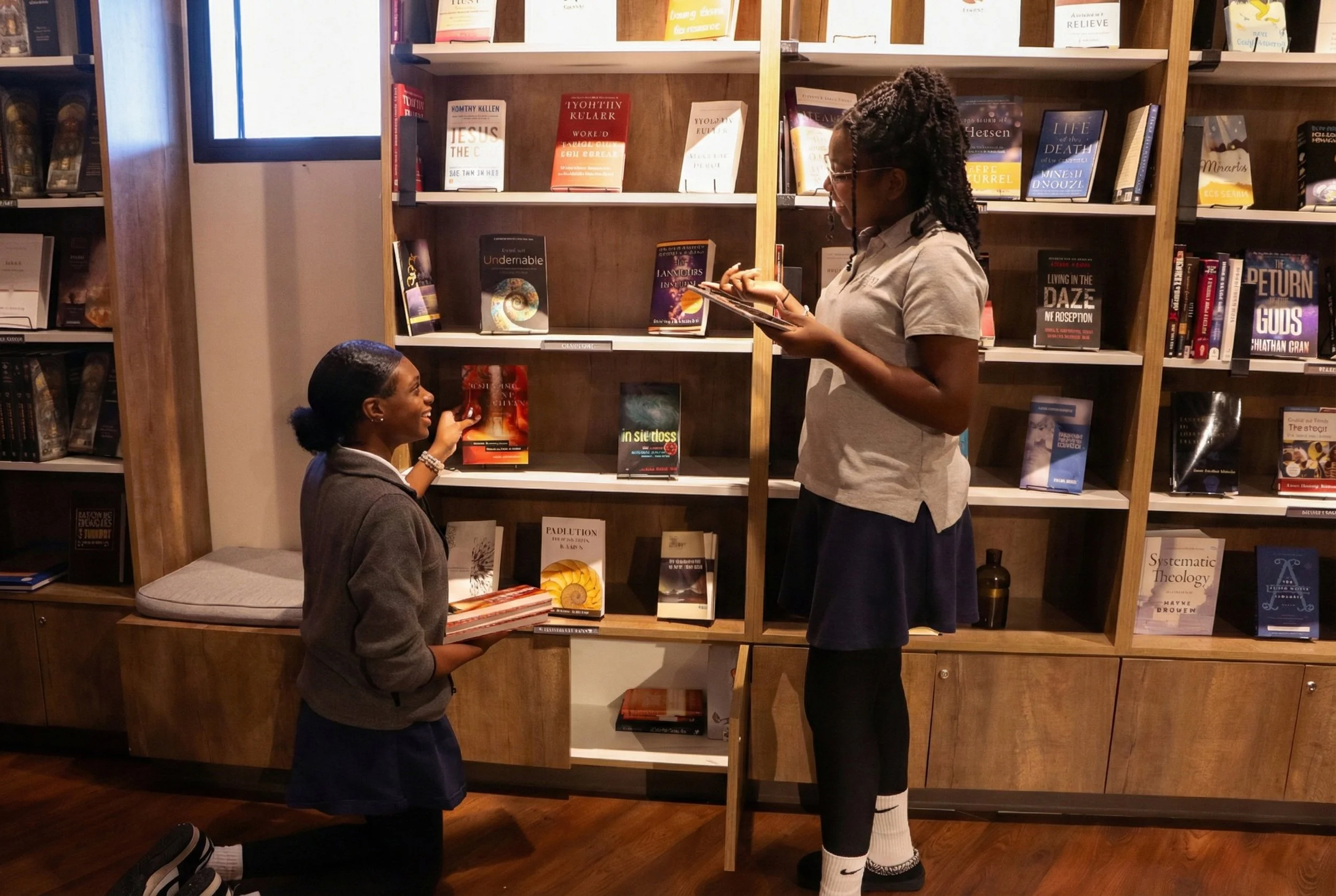 Two women in a bookstore or library, one is kneeling and handing a book to the other who is standing and reading. They are in front of a wooden bookshelf filled with various books.