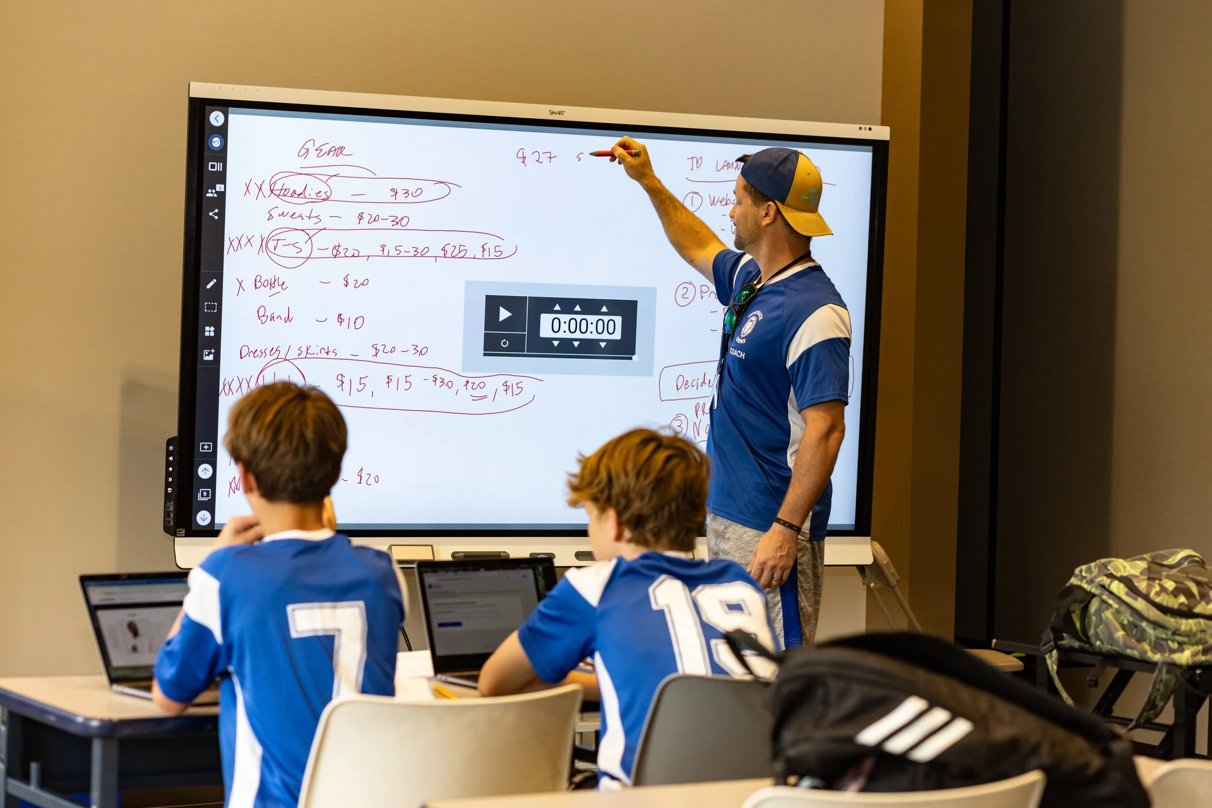 A teacher standing in front of a smartboard with handwritten notes, teaching a class of students wearing sports jerseys, with laptops open during a lesson.