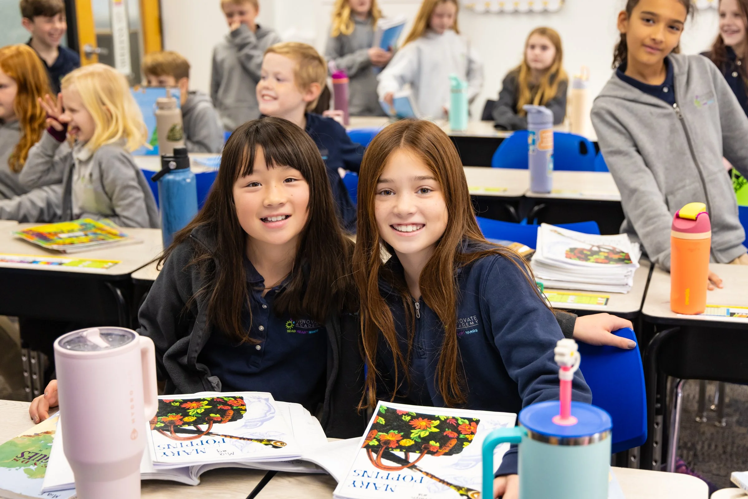 Two young girls sitting at a desk in a classroom, smiling at the camera, surrounded by other children, books, and water bottles.