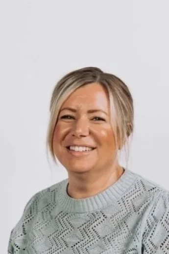 A woman with blonde hair smiling while sitting, wearing a light blue lace-patterned sweater and white pants, against a plain white background.