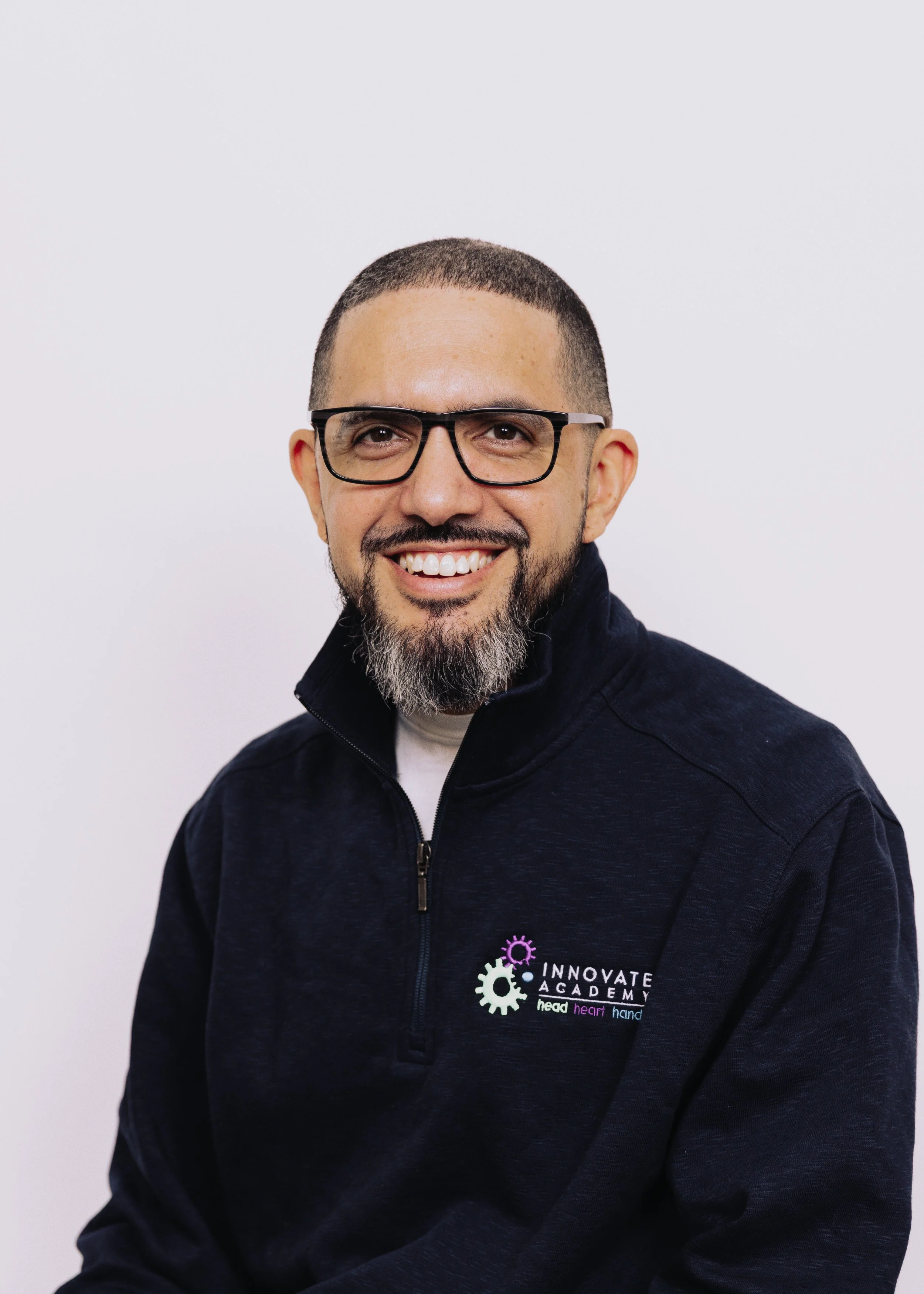 A man with glasses, a goatee, and a smile, wearing a black jacket with the logo 'Innovate Academy' on it, standing against a plain white background.