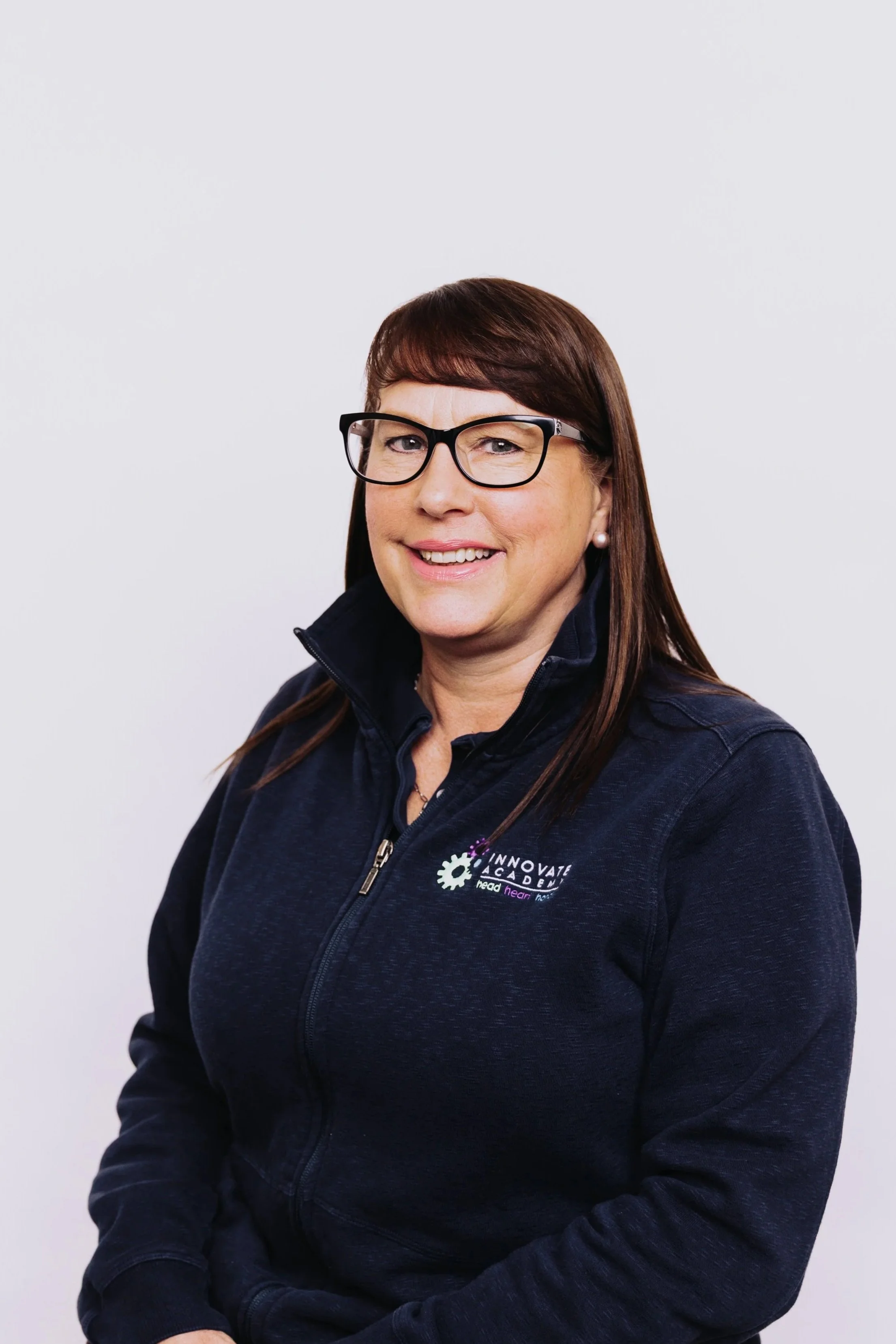 A woman with long brown hair, glasses, and silver earrings, wearing a black zip-up fleece jacket with a logo that reads 'Innovate Academy' on the left side, posing against a plain white background.