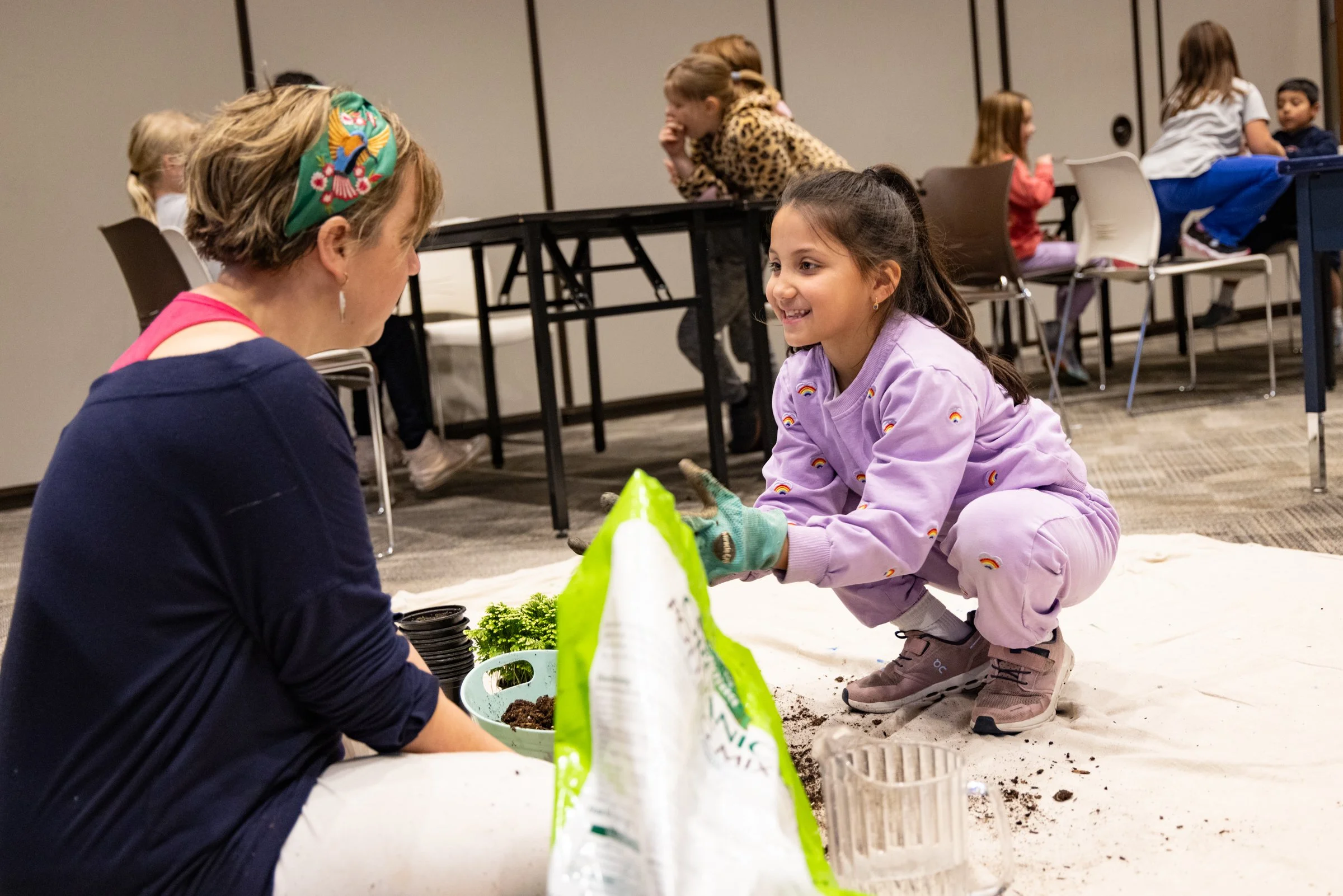 A young girl wearing a lavender sweatshirt and gardening gloves kneels on the floor, smiling and planting something in soil, while an adult woman with a colorful headscarf observes and assists her. There are other children in the background seated at tables in a classroom or activity space.