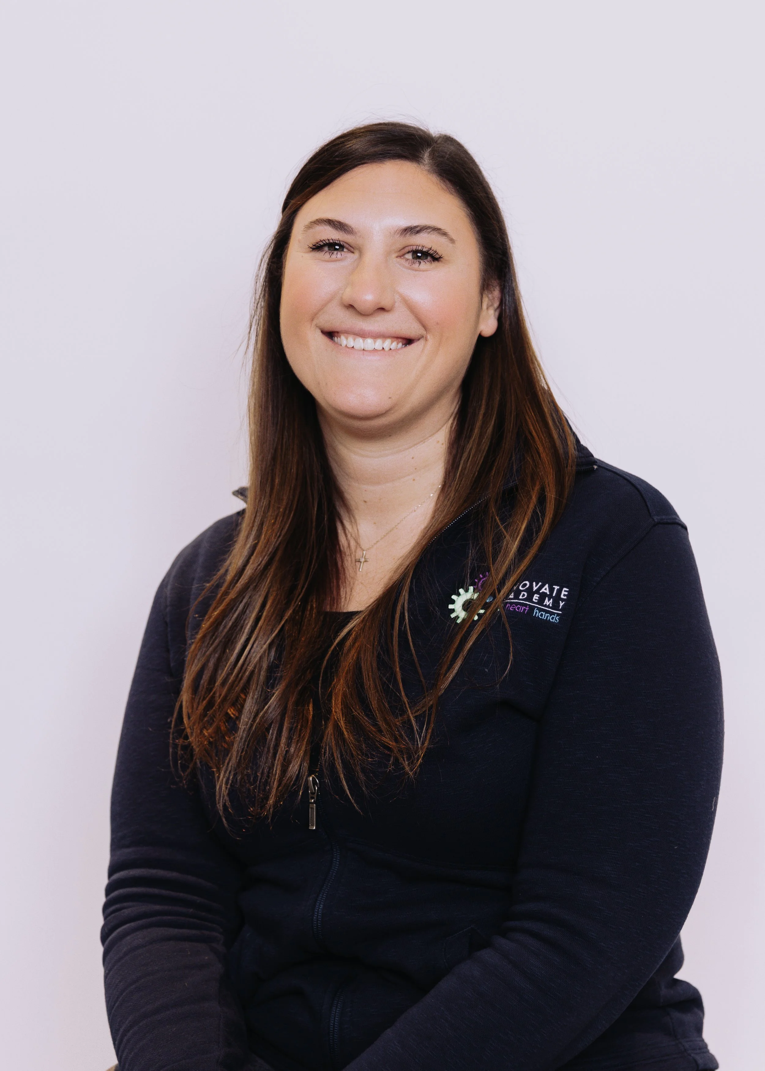 A woman with long brown hair smiling, wearing a black zip-up jacket with a badge that says "Innovate" and has a logo, against a plain light background.
